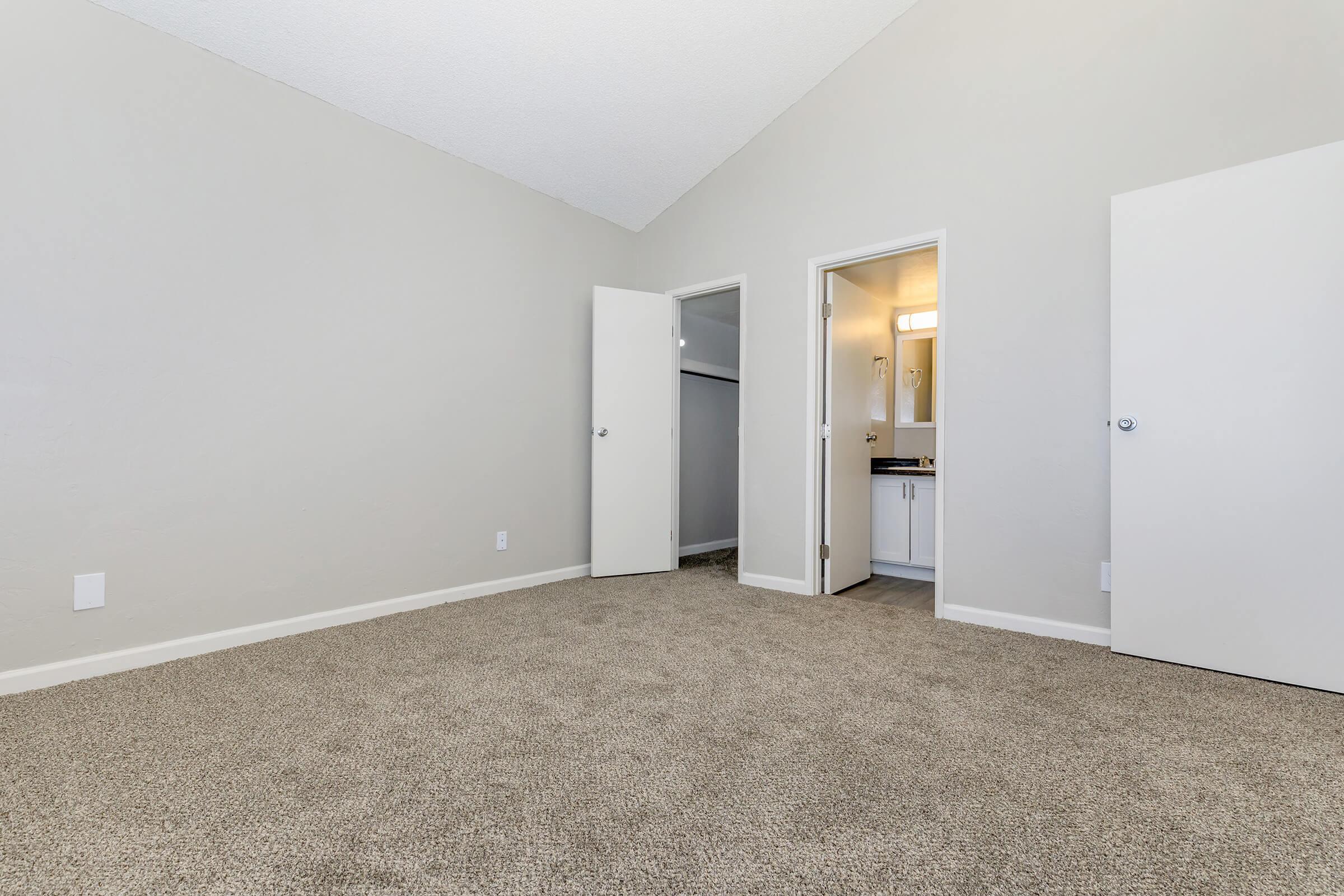 A spacious, empty room with beige carpet and light gray walls. There are two open doors leading to a closet and a bathroom. The ceiling has a slight slope, and the room is well-lit, suggesting a welcoming atmosphere.