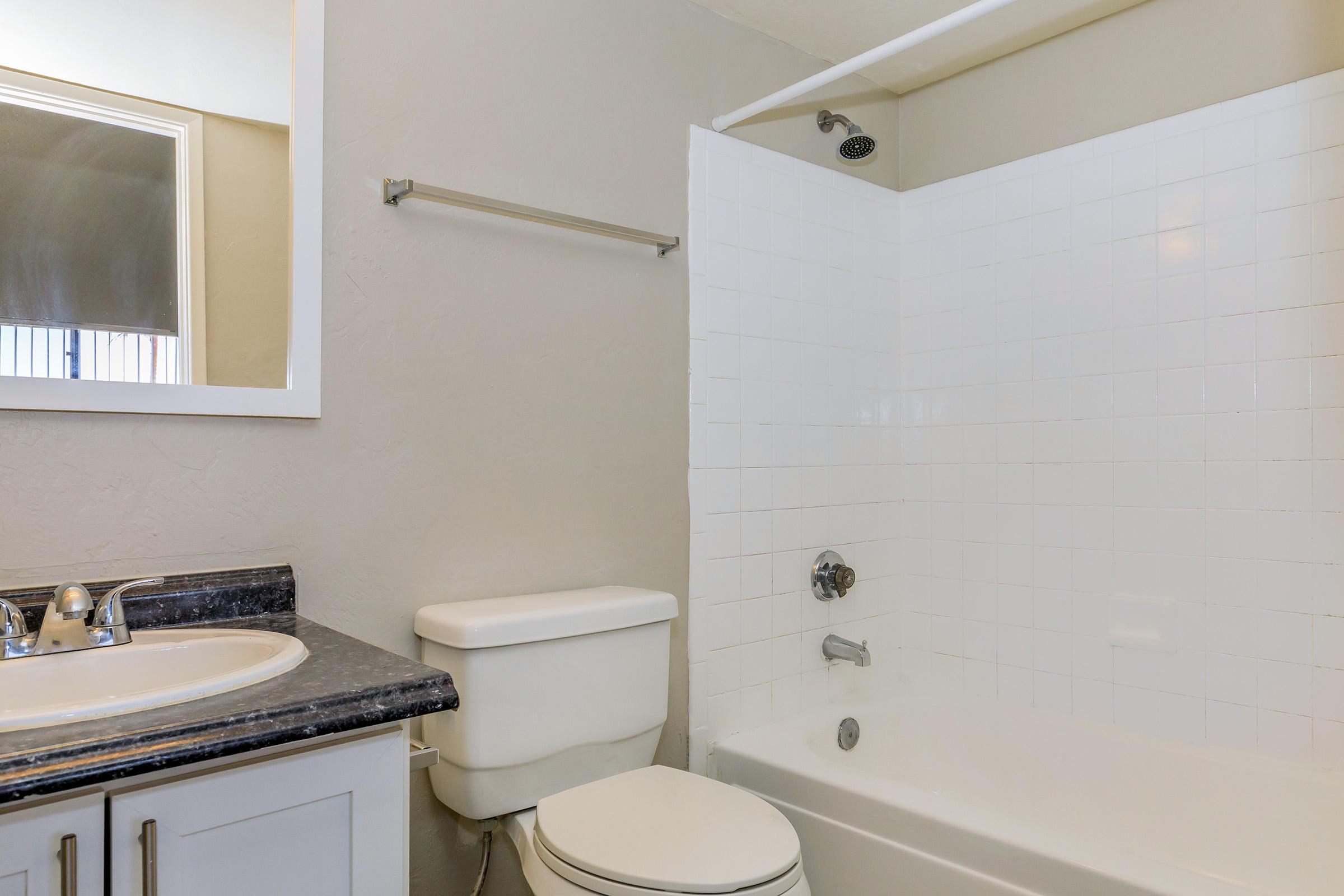 A clean, modern bathroom featuring a white bathtub with a showerhead, a white toilet, and a granite countertop with a sink. A mirror is mounted on the wall, and the walls are painted a light gray color. The space is well-lit and appears tidy.