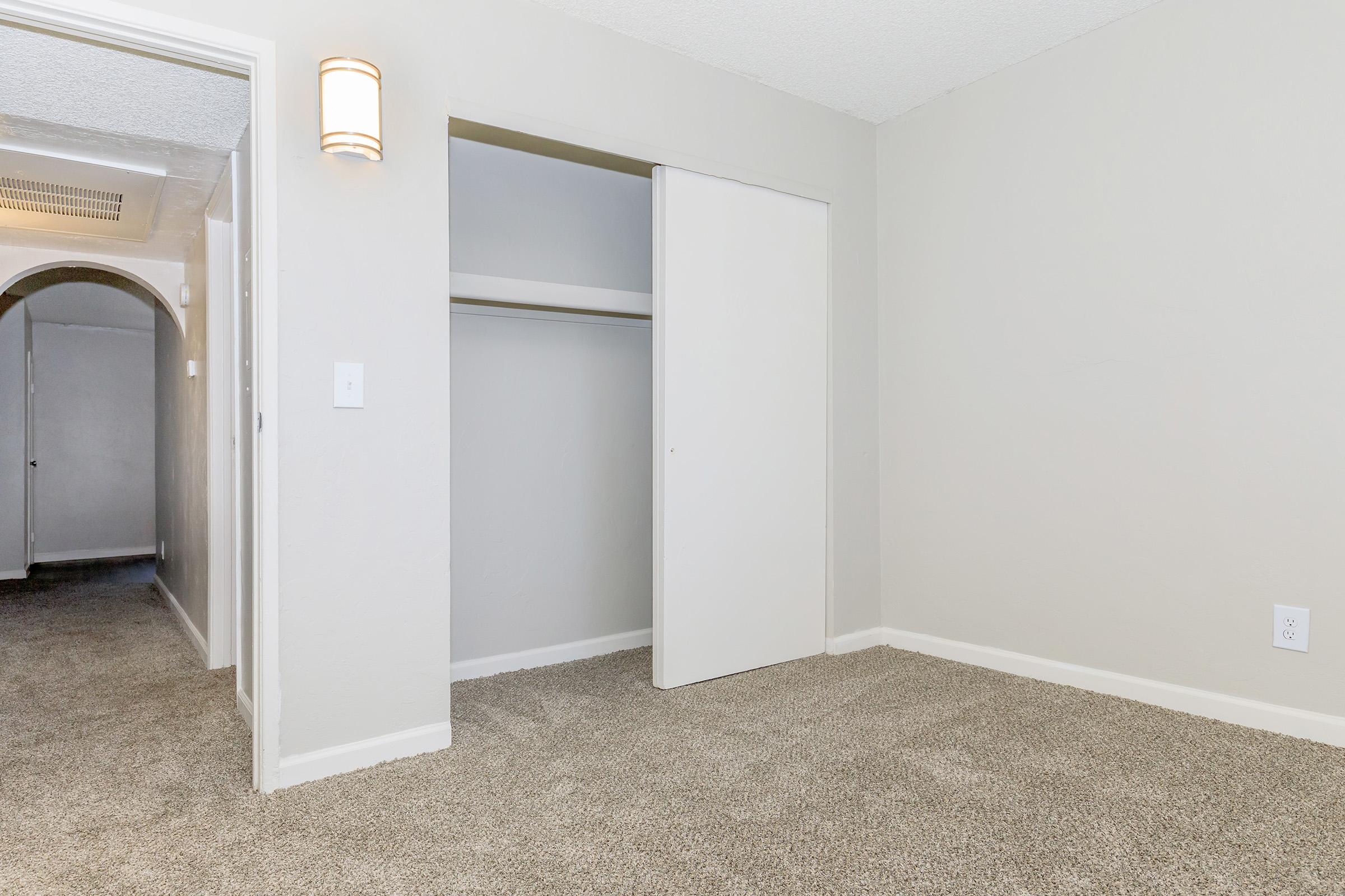Interior view of a light-colored room featuring a carpeted floor, a closed closet with sliding doors, and a wall-mounted light fixture. An arched doorway is visible in the background, leading to a hallway. The walls are painted a soft neutral shade, creating a spacious and inviting atmosphere.