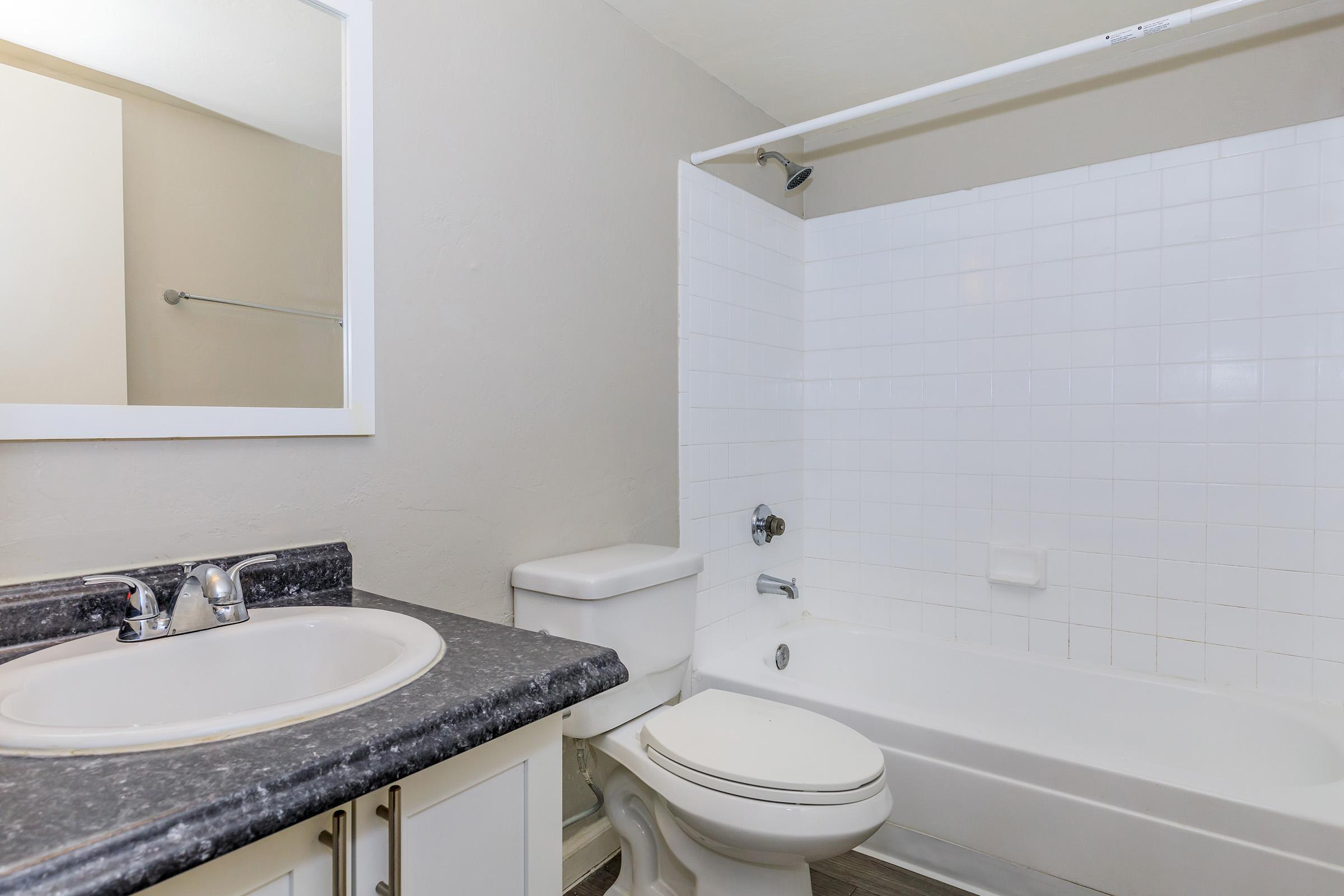 A clean bathroom featuring a white tiled bathtub with a shower, a modern toilet, and a sink with a dark countertop. The walls are painted in a light neutral color, and there is a simple framed mirror above the sink. The overall ambiance is bright and spacious.