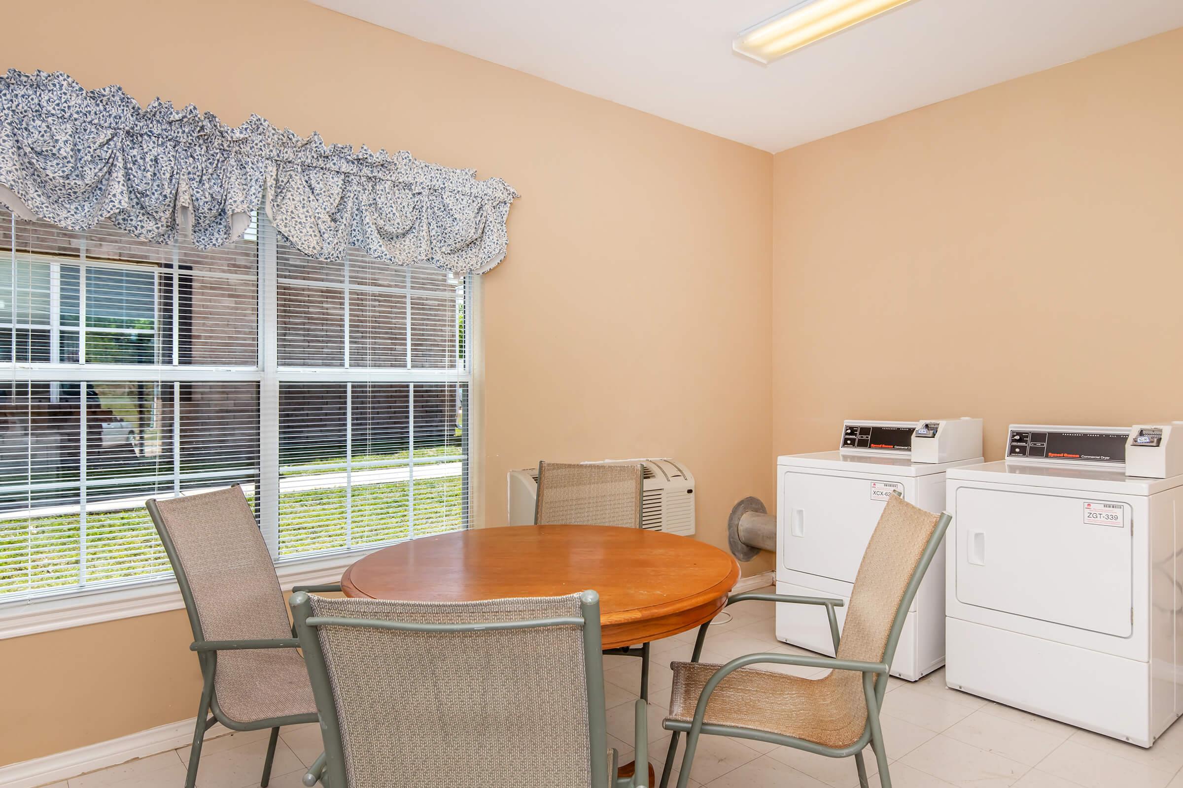 A bright laundry room featuring a round wooden table with four chairs, a window with light blue curtains, and two white washing machines side by side. The walls are painted a soft beige, and there is an air conditioning unit positioned against the wall.