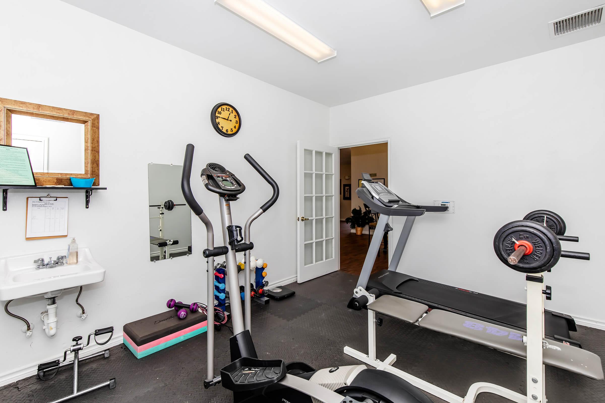 A home gym featuring an elliptical machine, treadmill, and weight bench with weights. There are colorful dumbbells on the floor, a wall clock, a mirror, and a small sink. The room has neutral-colored walls and is well-lit with overhead lighting. A door leads to another room.