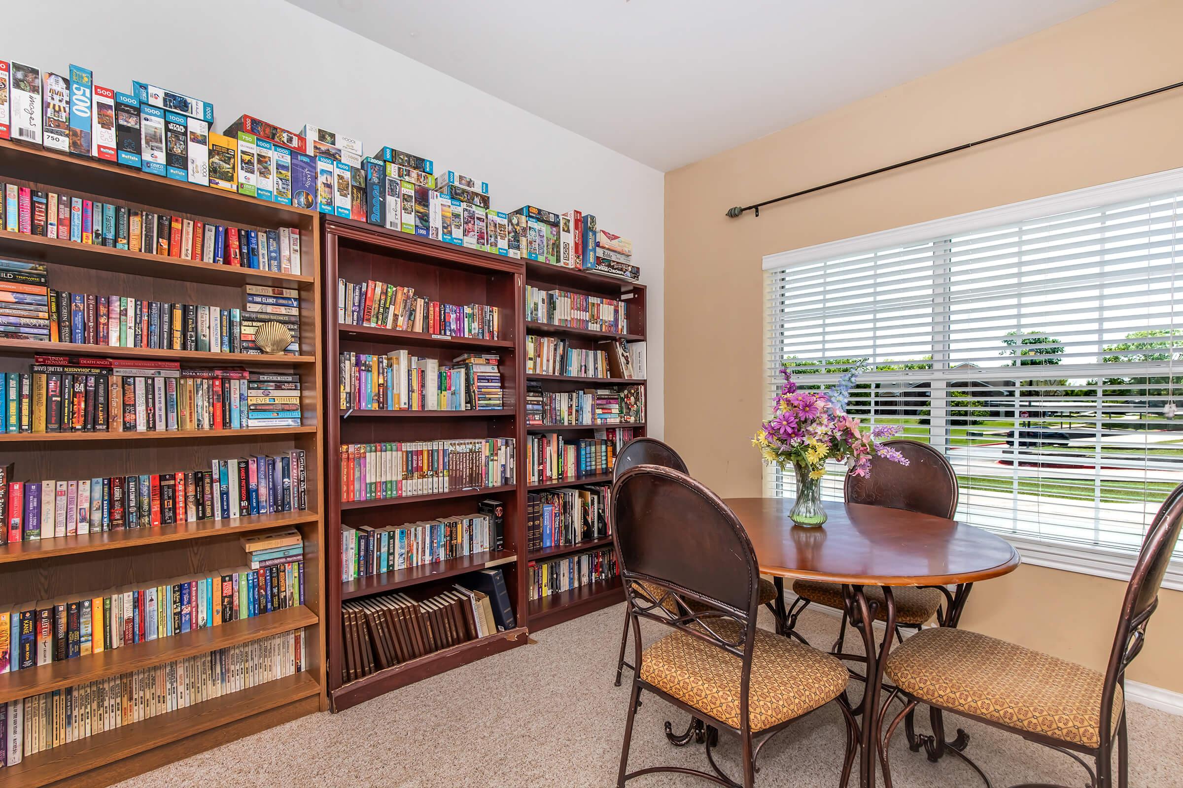A cozy reading nook featuring two wooden bookshelves filled with books, a round dining table with four chairs, and a vase of fresh flowers. Natural light streams in through a window with blinds, illuminating the space and providing a view of greenery outside.