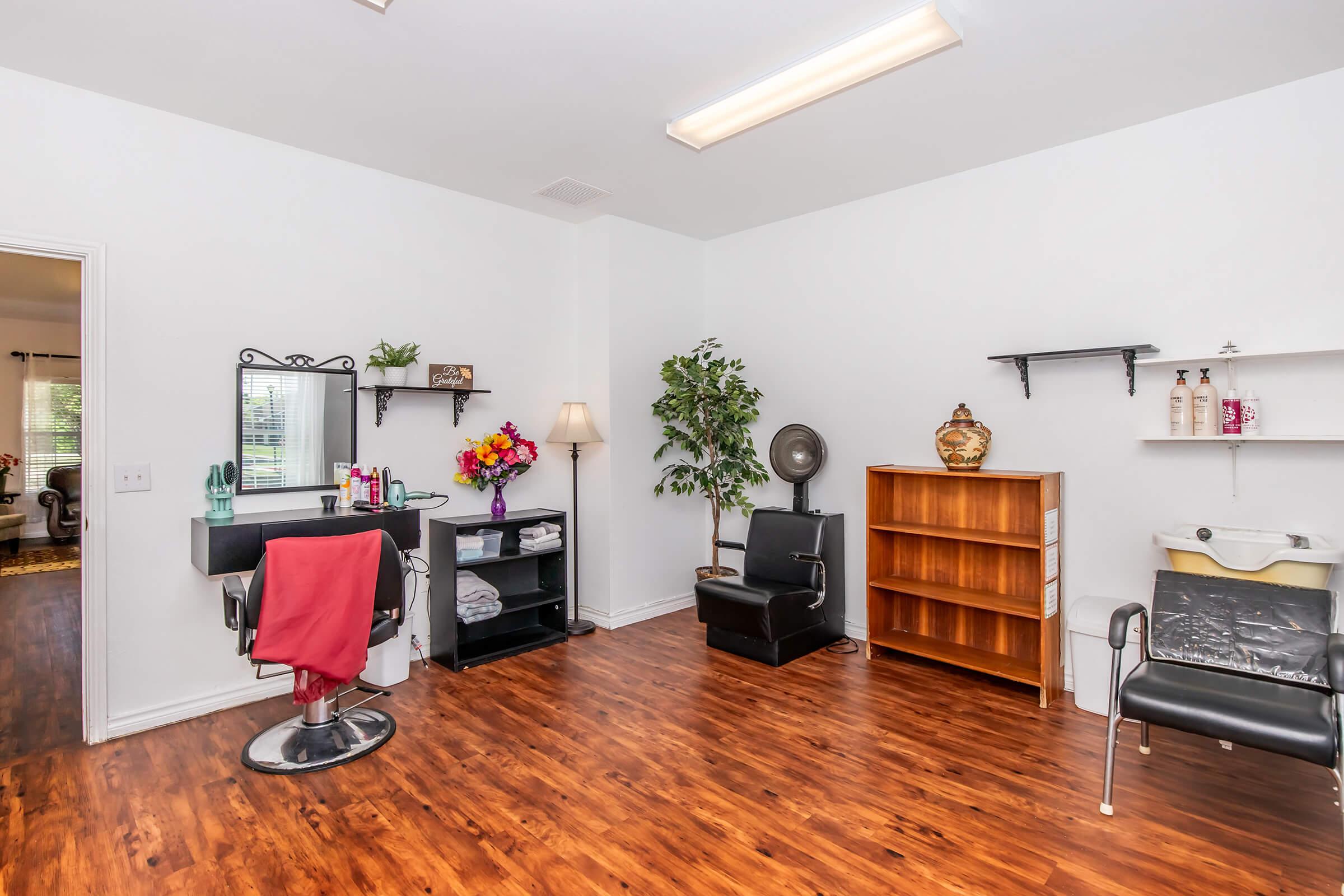 A bright and spacious hair salon interior featuring a styling station with a mirror and black salon chair, a shelf with hair products, a lamp, a potted plant, a wooden bookshelf, a chair, and a wash station. The walls are painted white and the floor is a warm wood tone, creating a welcoming atmosphere.