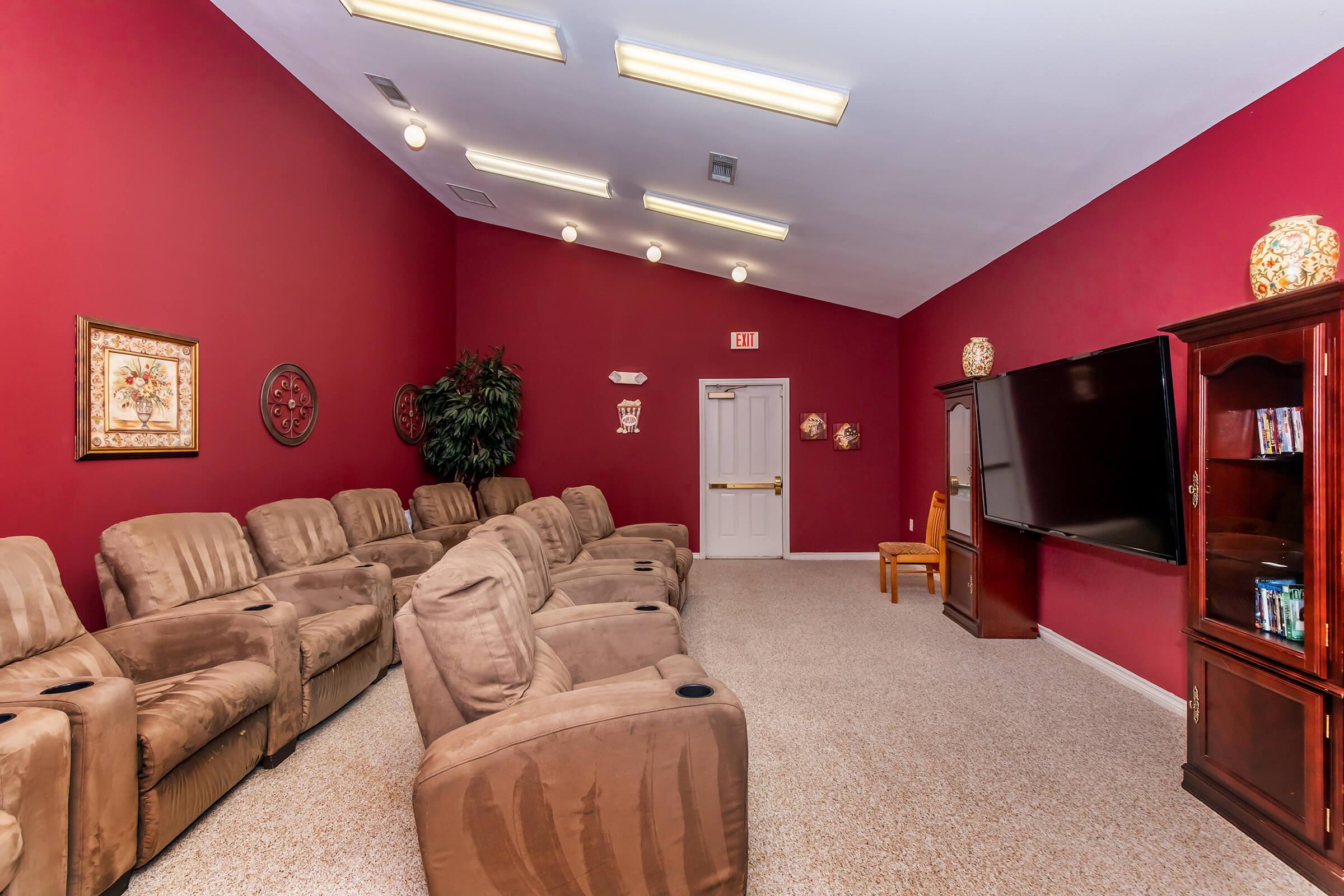 Cozy home theater room with red walls, featuring plush reclining seats, a large flat-screen TV, and decorative wall art. The space has warm lighting and a welcoming atmosphere, ideal for movie nights or relaxation. A wooden chair and cabinet add to the room's functionality.