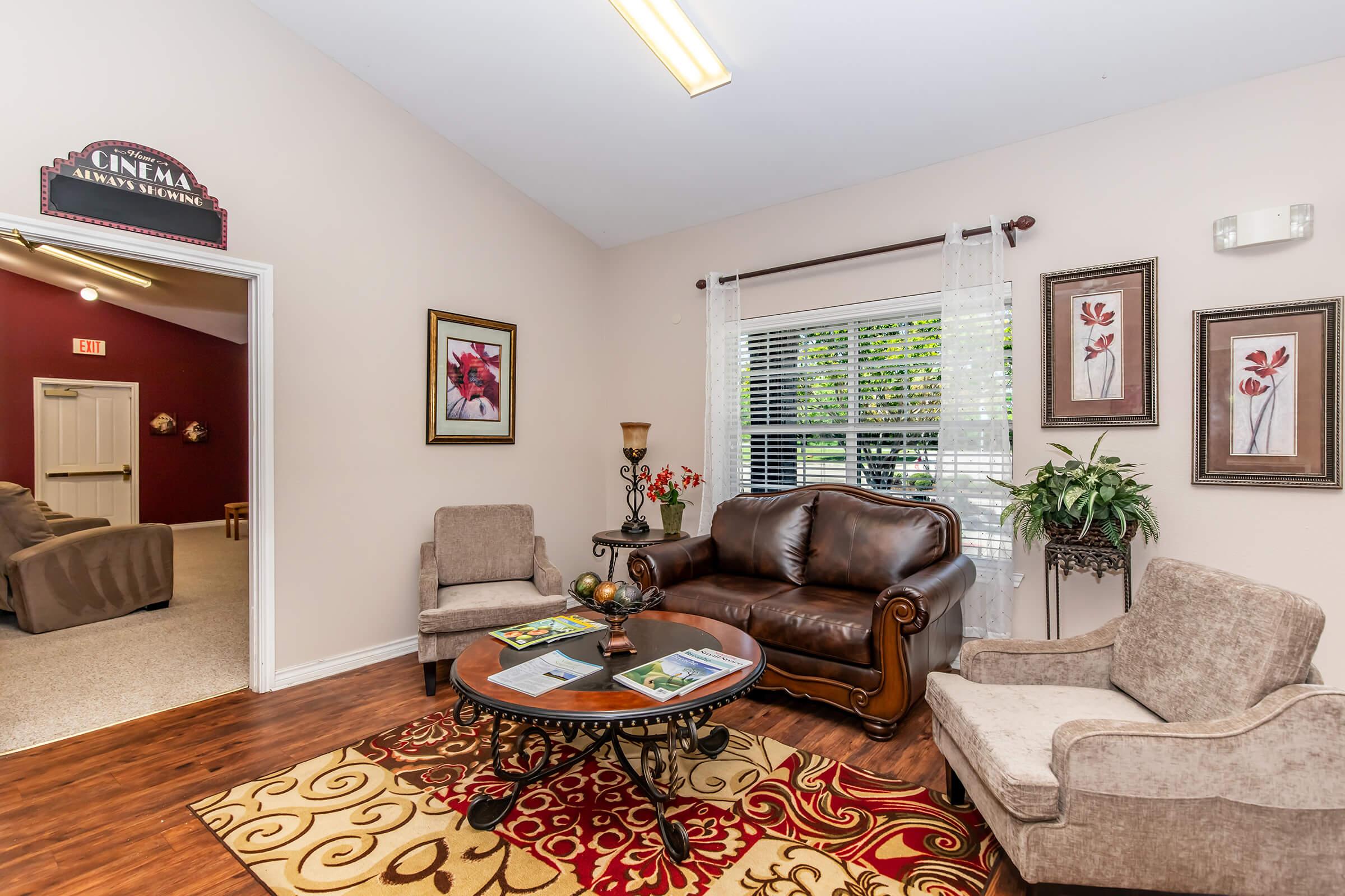 A cozy sitting area featuring a brown leather sofa and two armchairs, surrounded by decorative plants and framed artwork. A circular coffee table displays magazines. The room is well-lit with natural light coming through a window, and there's a sign labeled "Cinema" on the wall.