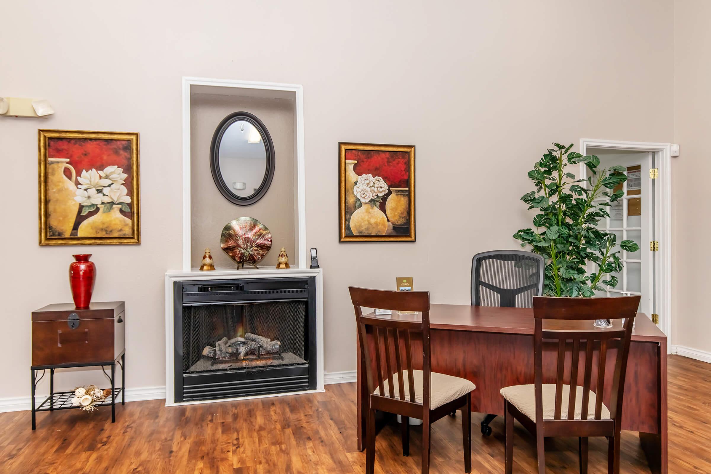A cozy office space featuring a wooden desk with a black chair, two decorative paintings on the walls, a small cabinet, an electric fireplace, and a potted plant. The flooring is a warm wood tone, creating an inviting atmosphere.
