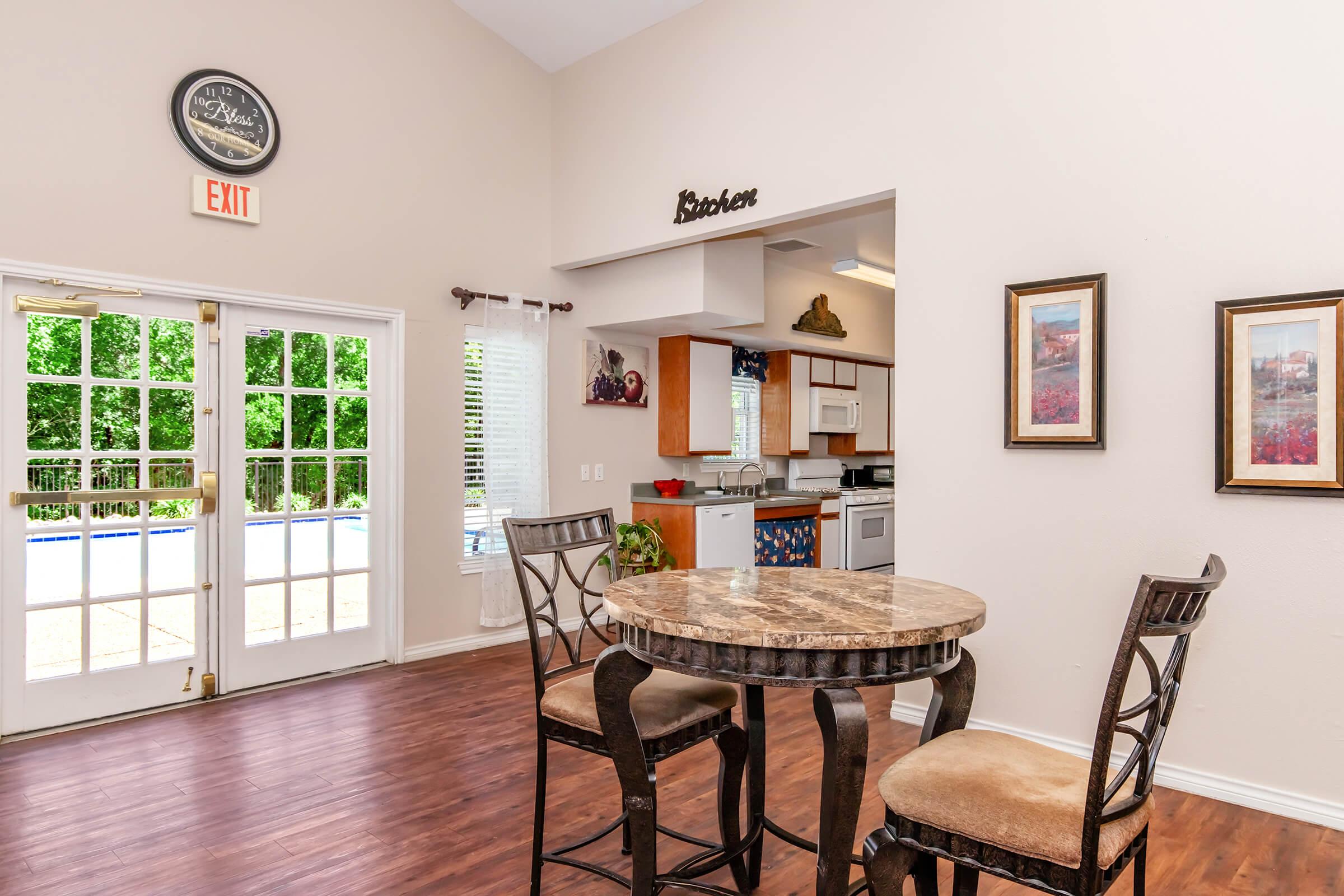 A cozy dining area featuring a round marble table with two chairs. In the background, there's a kitchen area with cabinets and a decorative piece. Large double doors lead outside, letting in natural light. The walls are painted in neutral tones, and framed artwork adds a touch of decor.