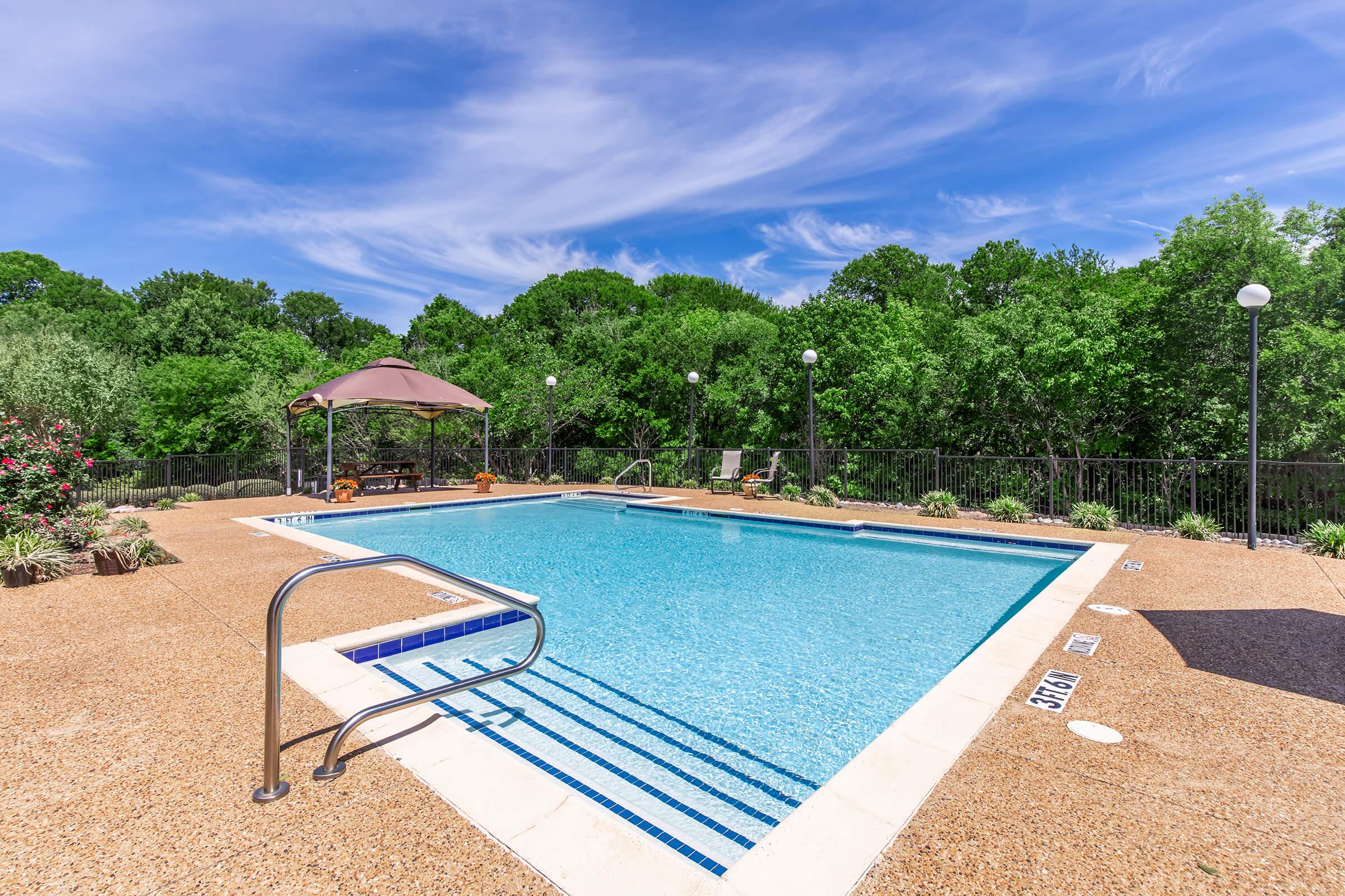 A clear swimming pool surrounded by a patio with lounge chairs and a gazebo. Lush green trees and a blue sky with scattered clouds are visible in the background, creating a serene outdoor setting perfect for relaxation and leisure.