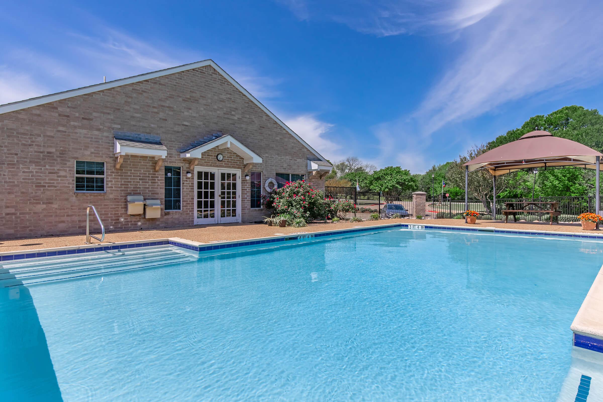 A clear blue swimming pool in front of a brick building, surrounded by greenery. The pool has a shallow entry with steps and is bordered by a stone deck. In the background, a gazebo with a table can be seen, along with flowering plants adding color to the scene. A bright sky with a few clouds completes the peaceful atmosphere.