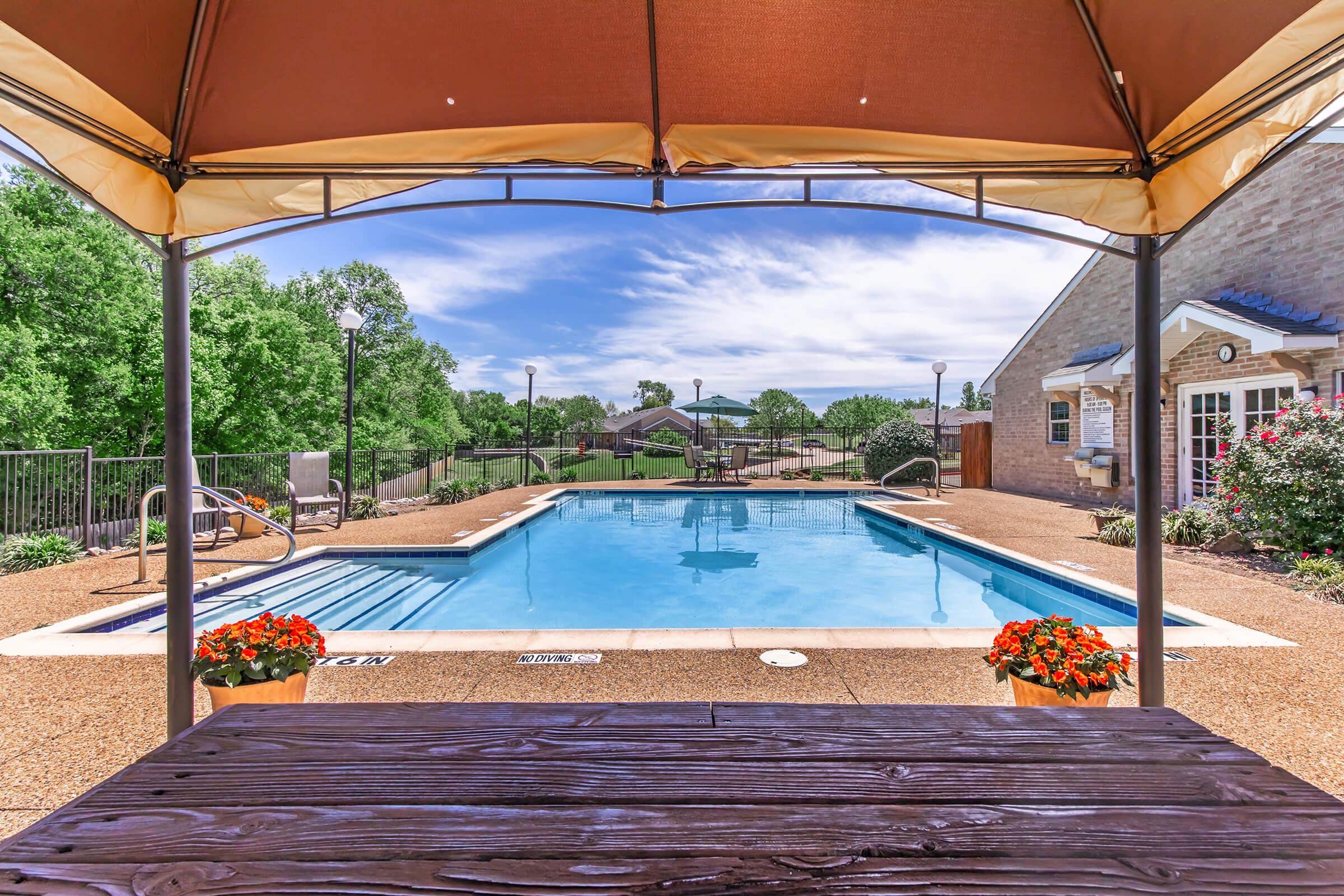 View of a swimming pool from under a shaded gazebo. The pool is blue and surrounded by a patio. In the foreground, there are flower pots with vibrant flowers. In the background, green trees and a grassy area can be seen, along with a building and pathways, all under a clear blue sky with clouds.