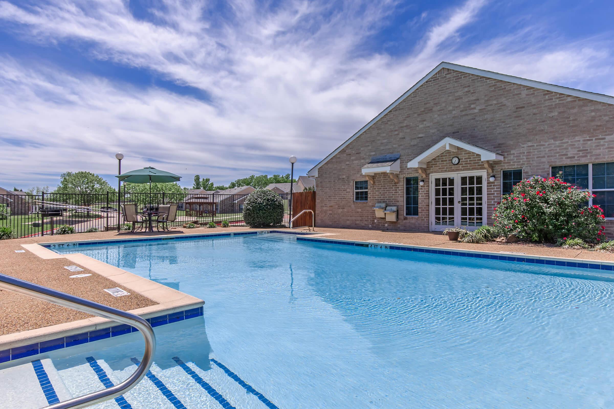 A clear blue swimming pool surrounded by a well-maintained area featuring lounge chairs, an umbrella, and greenery. In the background, there is a brick building with large windows and a fenced area. The sky is partly cloudy, creating a serene atmosphere.