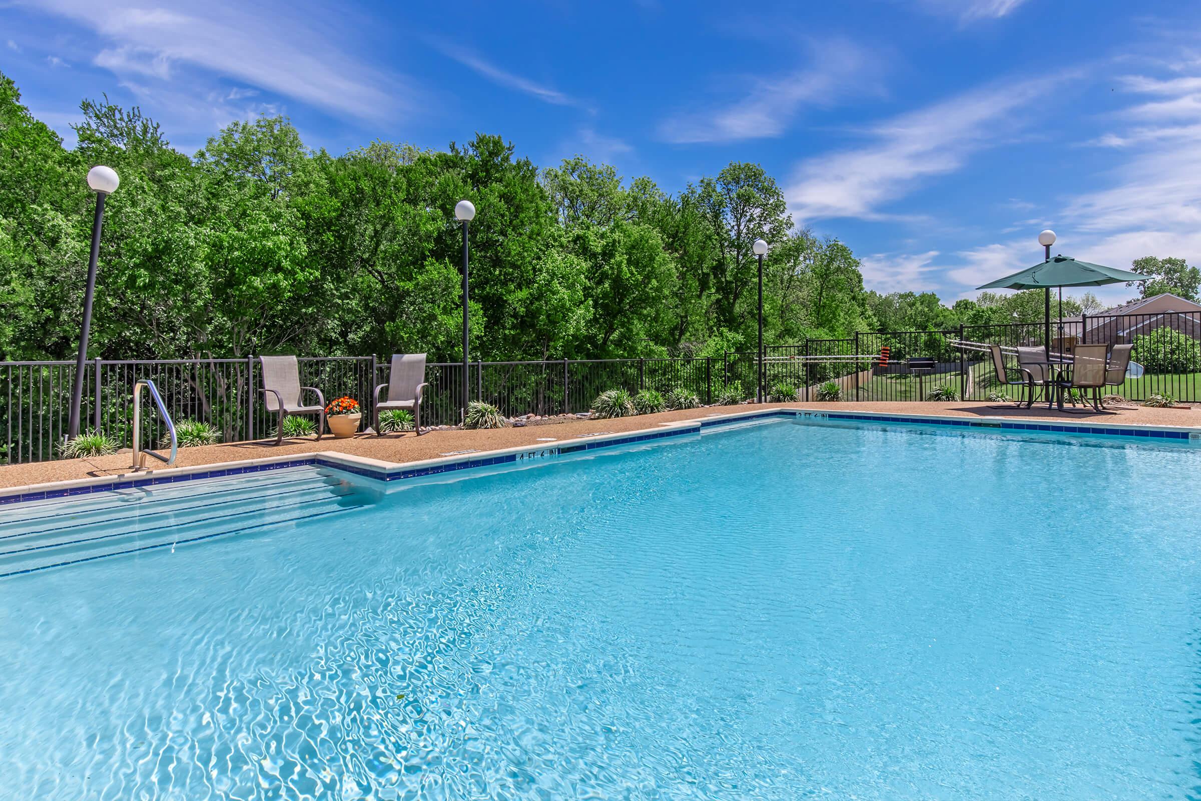 A clear blue swimming pool surrounded by a deck, featuring lounge chairs and a table with an umbrella. Lush green trees are visible in the background under a bright blue sky with scattered clouds, creating a vibrant and inviting atmosphere.