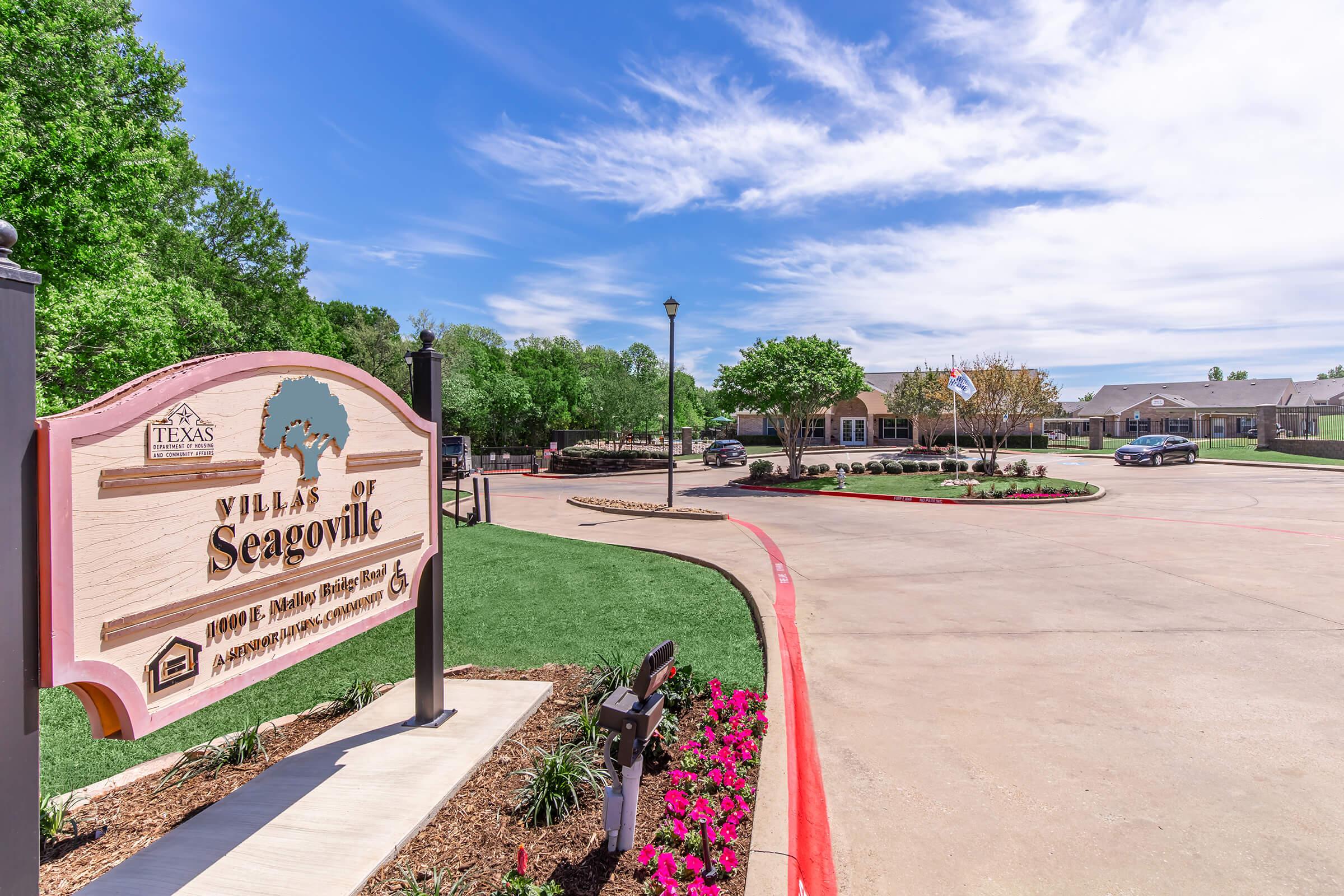 Sign for the Villas of Seagoville, located in Texas, with landscaped gardens and flower beds in the foreground. The entrance features a well-maintained road and residential buildings visible in the background, showcasing a sunny day with green trees and blooming flowers.