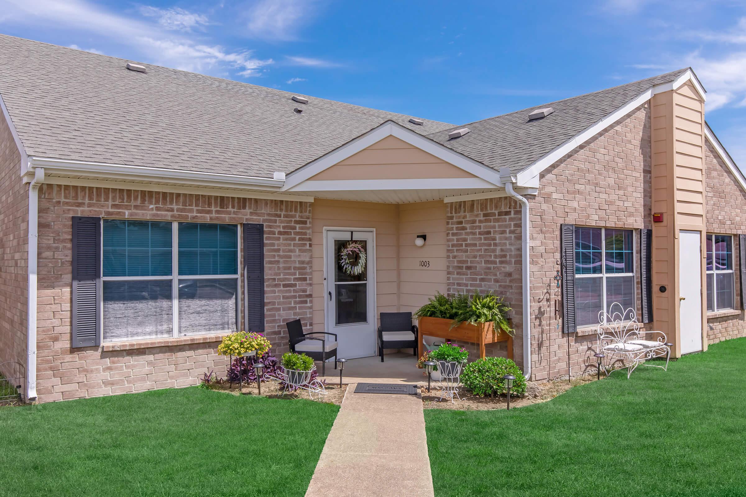 A charming brick house with a small porch, featuring a wreath on the front door, surrounded by greenery and decorative plants. The well-maintained lawn is presented with a stone pathway leading to the entrance. Clear blue sky enhances the inviting appearance of the home.