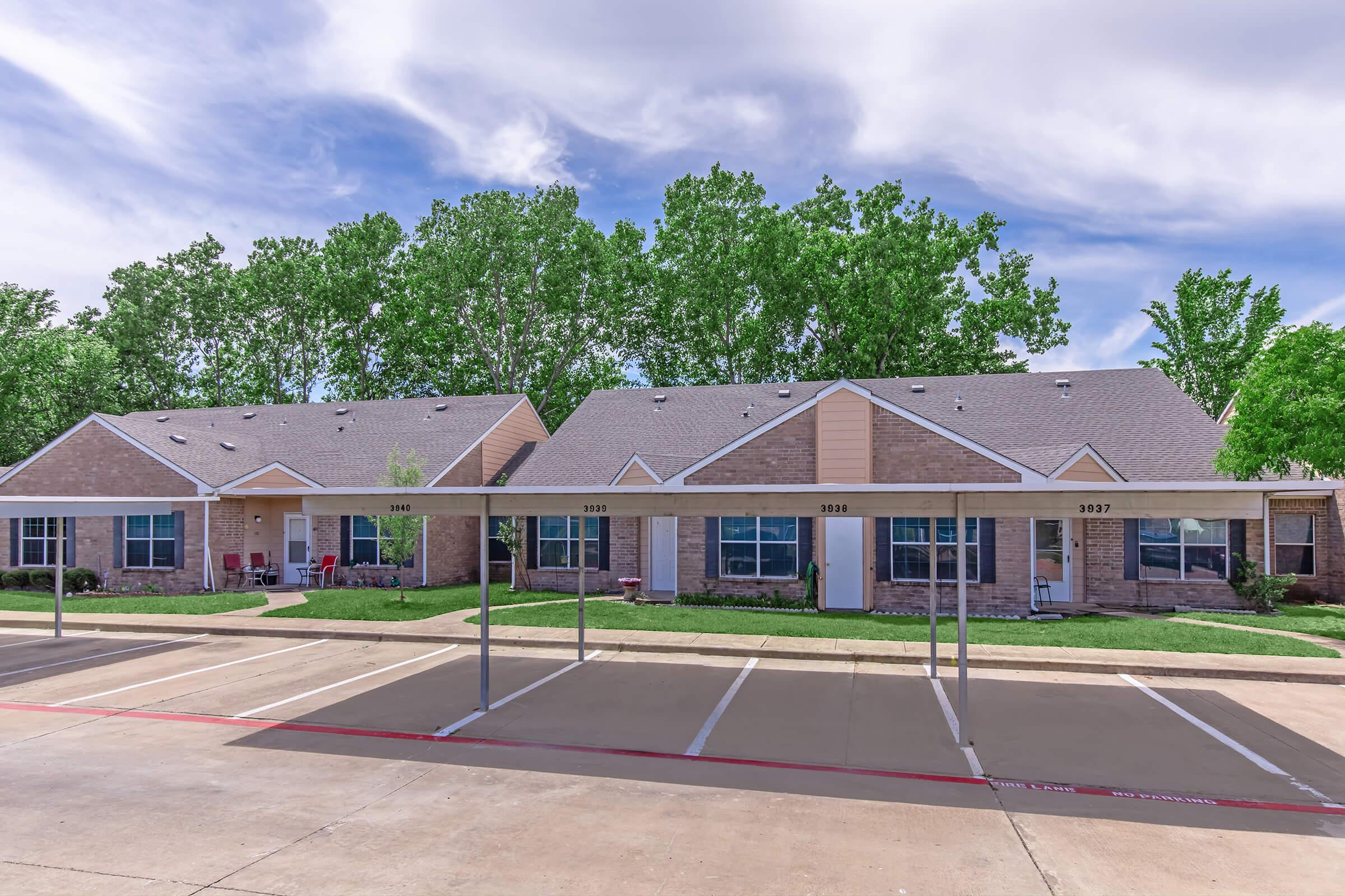A row of single-story residential units with a walkway in front, surrounded by green trees. The buildings have beige and brown exteriors, with parking spaces in front of them. The sky is partly cloudy, adding a calm atmosphere to the scene.