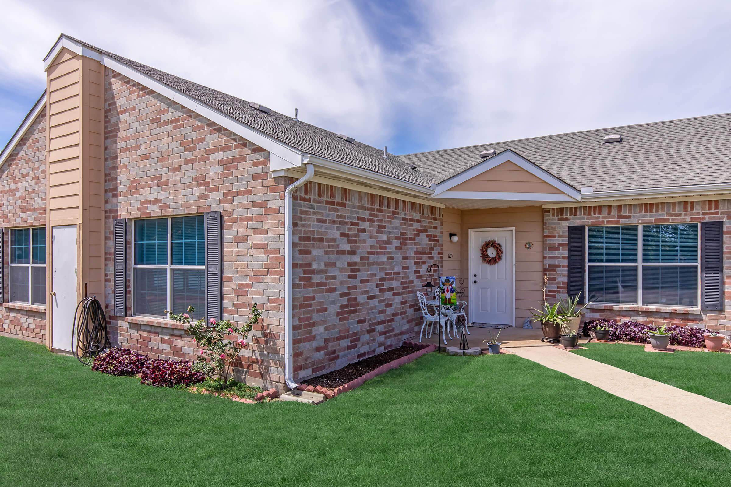 A well-maintained brick house with a light-colored siding, featuring a welcoming front porch with a wreath on the door. Lush green grass surrounds the walkway, and potted plants add color to the entrance. Windows are adorned with shutters, creating a charming and inviting atmosphere.