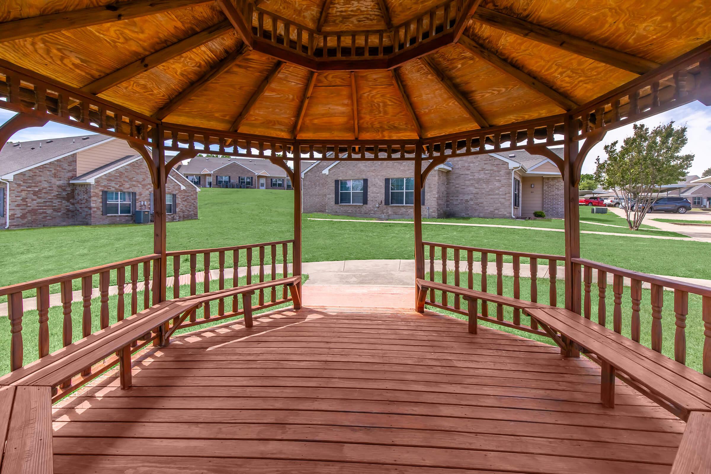 A wooden gazebo with a hexagonal roof and benches, set in a grassy area. The view from inside the gazebo shows nearby houses and a clear sky, creating a tranquil outdoor atmosphere.