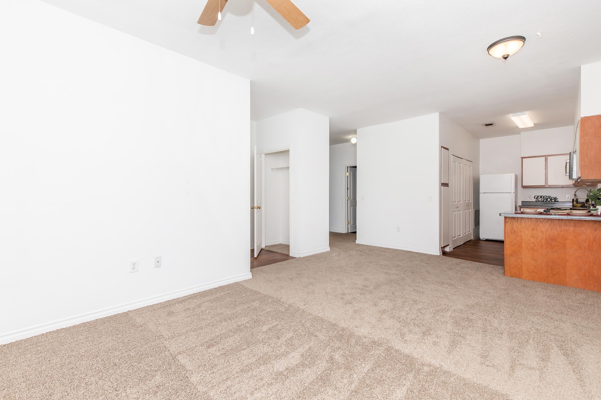A spacious, light-colored living area with beige carpet, a ceiling fan, and a doorway leading to an adjacent room. The kitchen is partially visible in the background, featuring wood cabinets and appliances. The walls are plain white, creating a bright and open atmosphere.