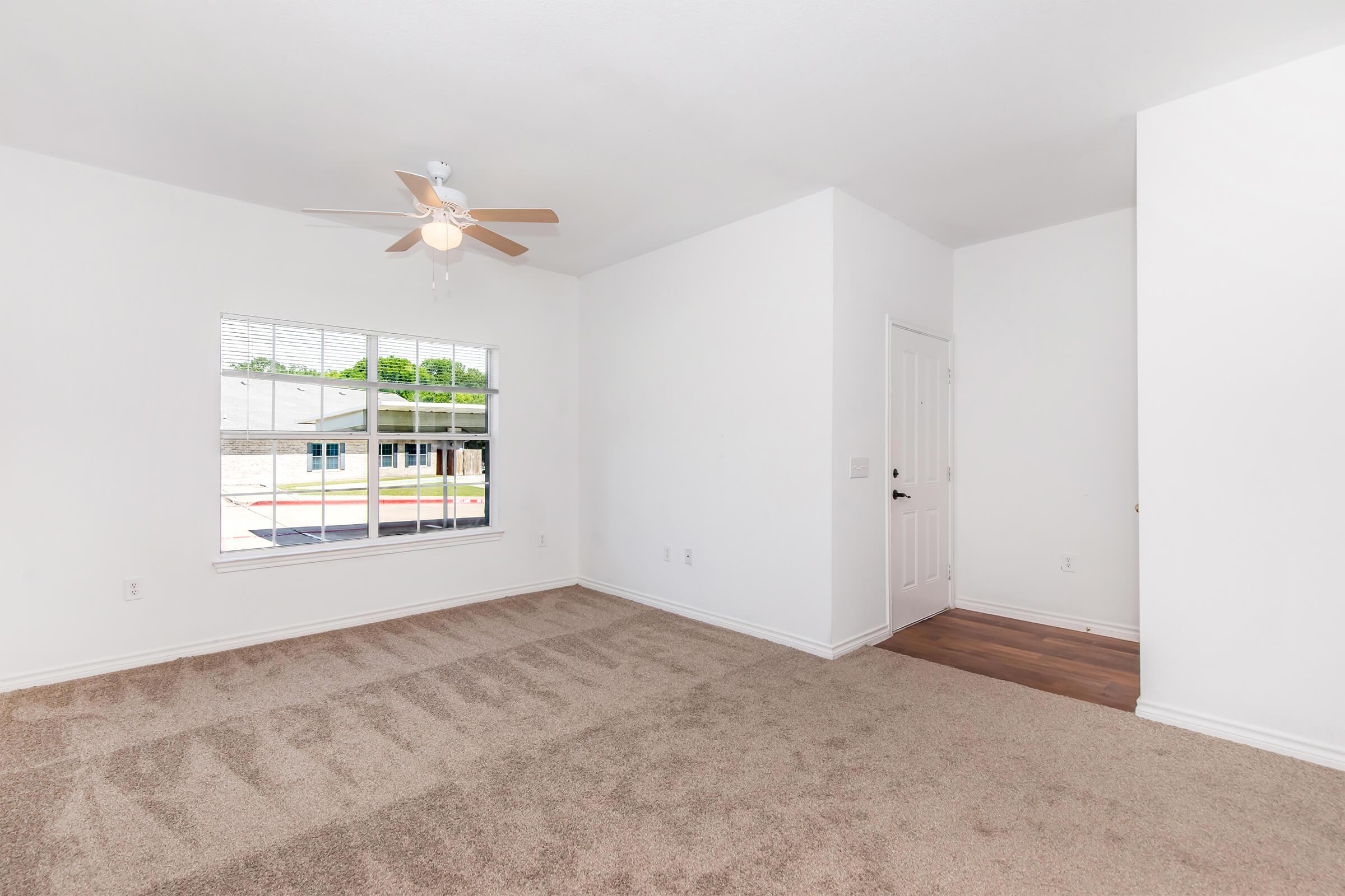 A bright, empty room featuring light-colored walls and carpet. There is a ceiling fan with wooden blades, a large window allowing natural light, and a door visible on the right. The space is uncluttered, making it suitable for various furniture arrangements.