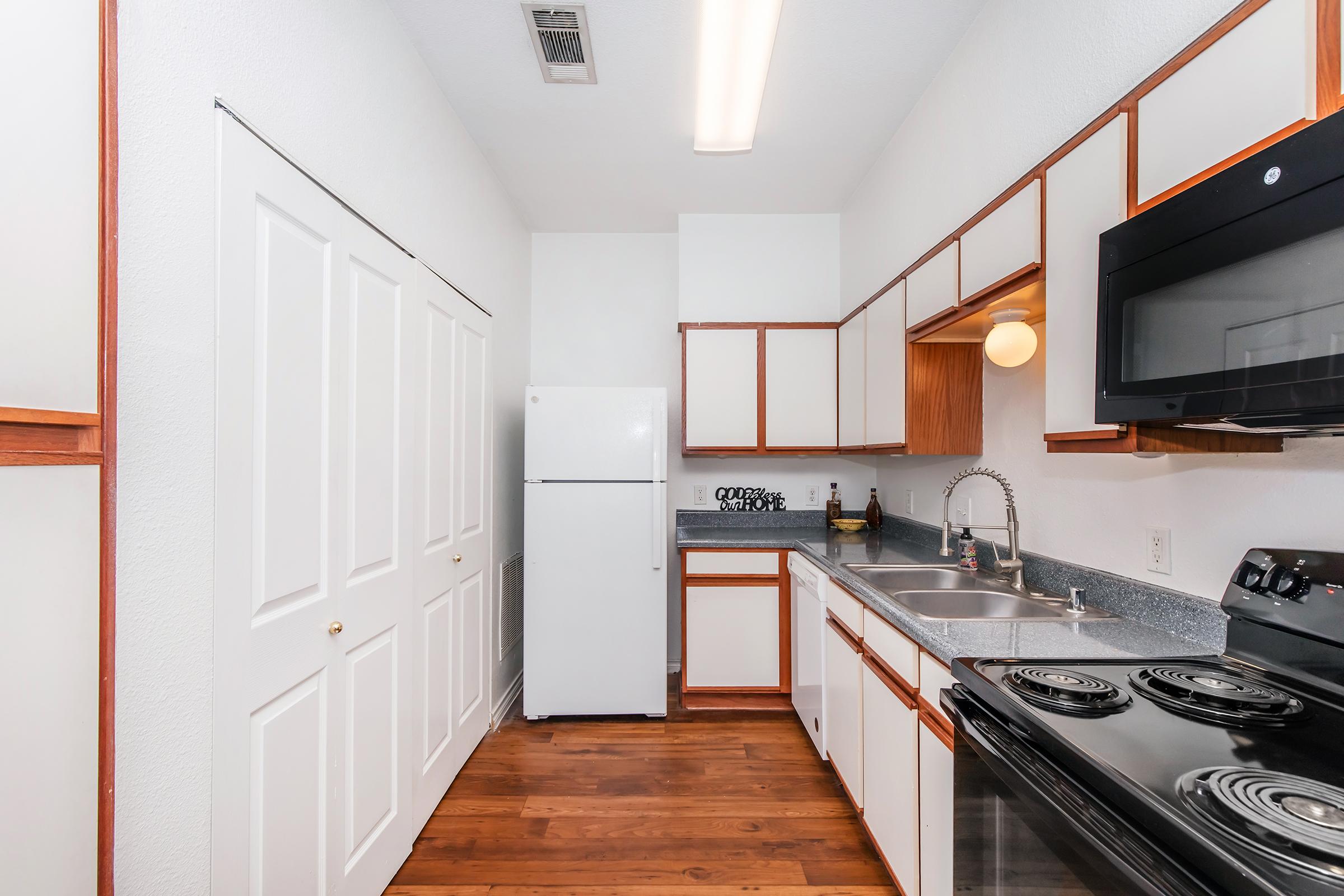 A modern kitchen featuring white cabinets with wooden accents, a gray countertop, a stainless steel sink, and black appliances, including an electric stove and microwave. The space has wooden flooring and ample storage, with a refrigerator and a light fixture overhead.