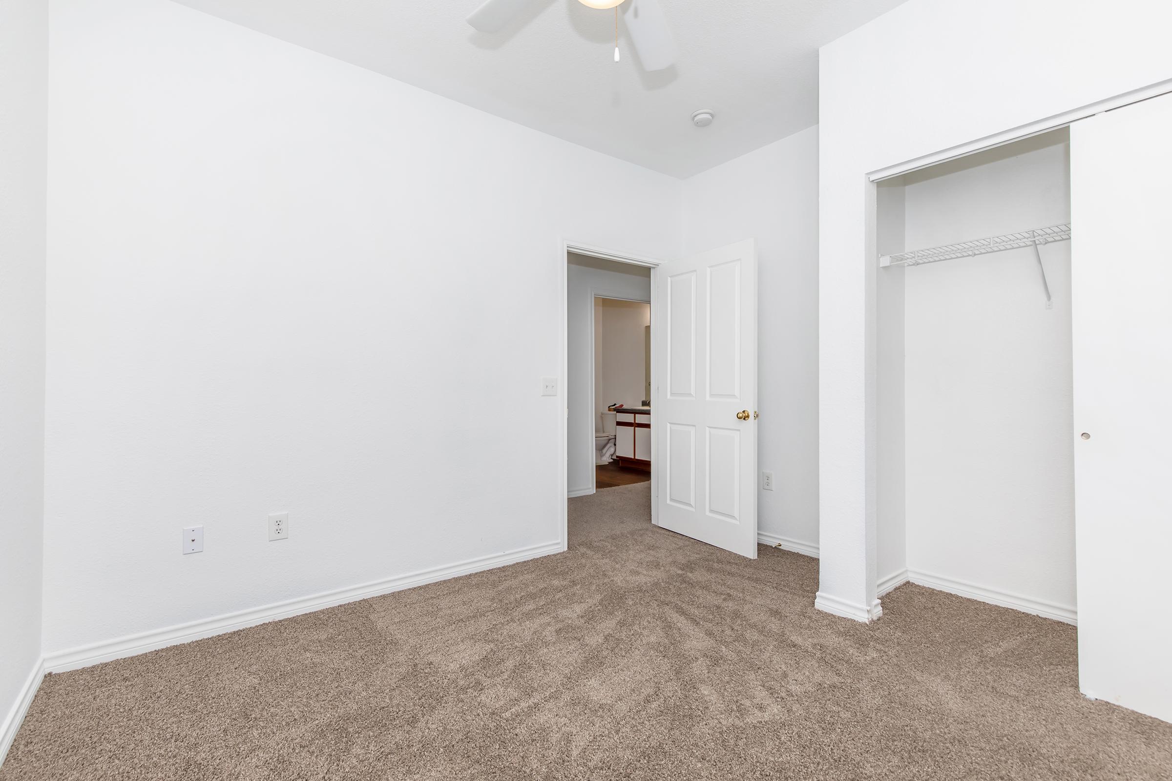A vacant room with white walls and carpeted flooring. There is an open door leading to another room, and a closet with sliding doors on the right side. The ceiling features a ceiling fan, and there is ample natural light illuminating the space, creating a clean and modern atmosphere.