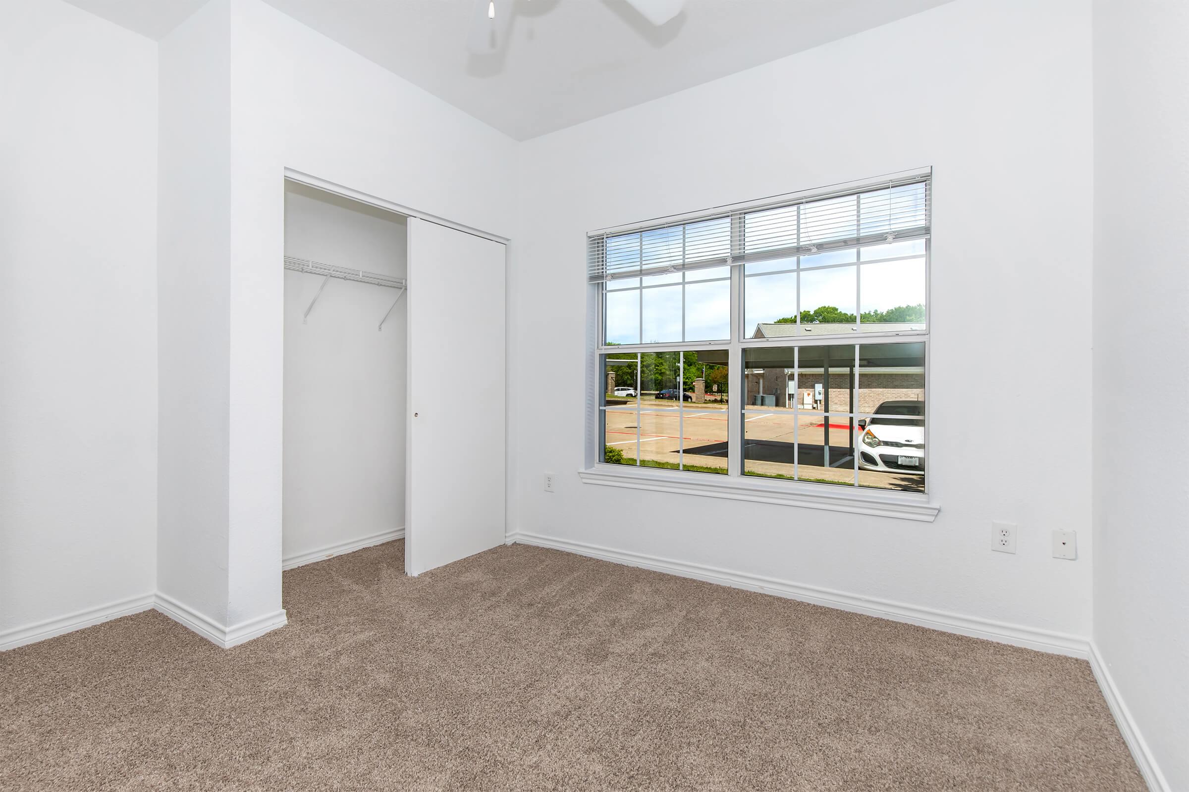 A clean, empty bedroom with light-colored walls and carpet. The room features a large window allowing natural light, showcasing a view outside, and a closet on one side. The space has a minimalist design, making it ready for furnishing and personalization.