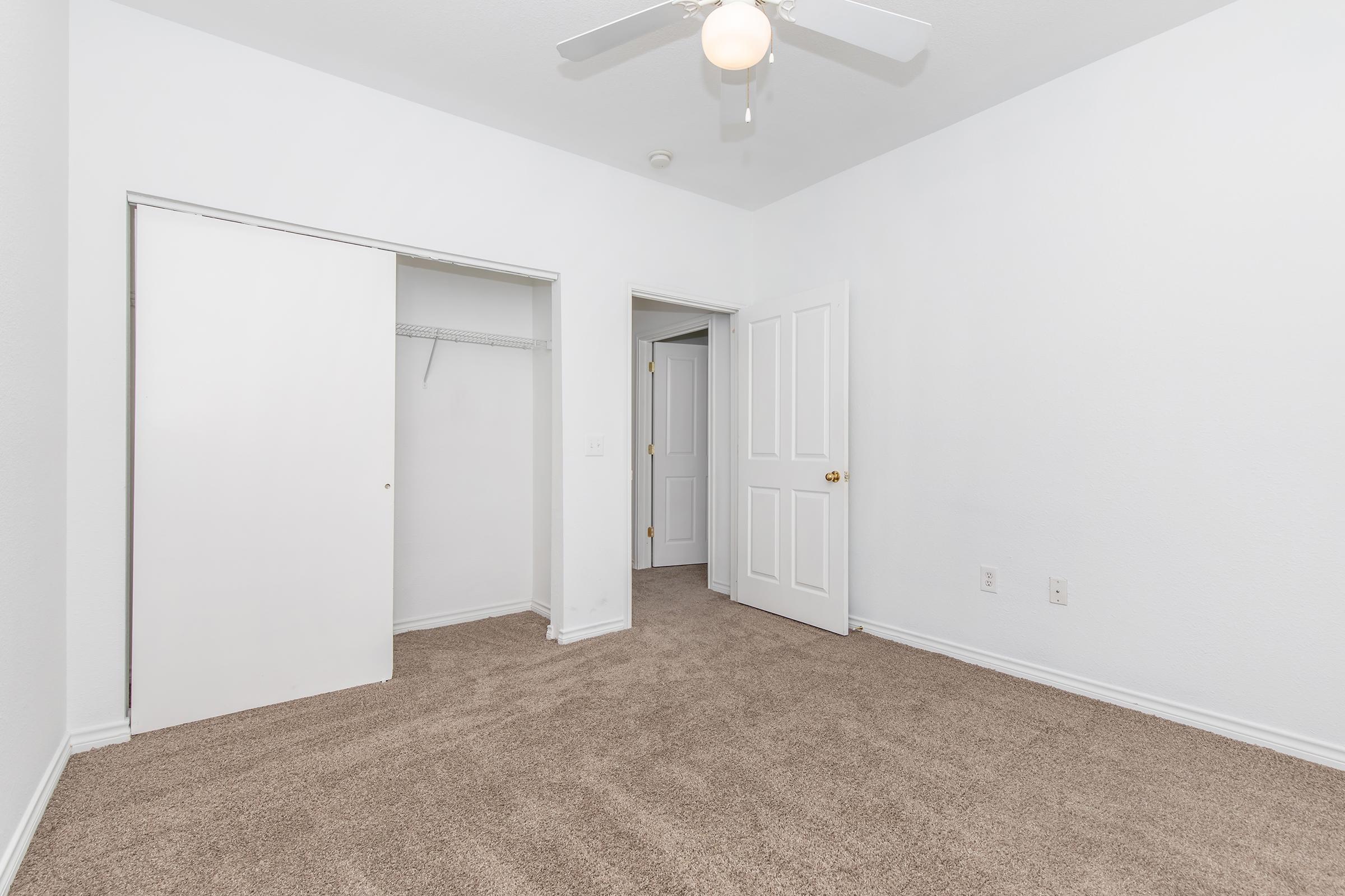 A clean, empty room with light beige carpet, white walls, and a ceiling fan. There are two doors: one leading to a closet and another to the hallway. The space is well-lit, giving a fresh and open feel, ideal for personal customization or moving in furniture.