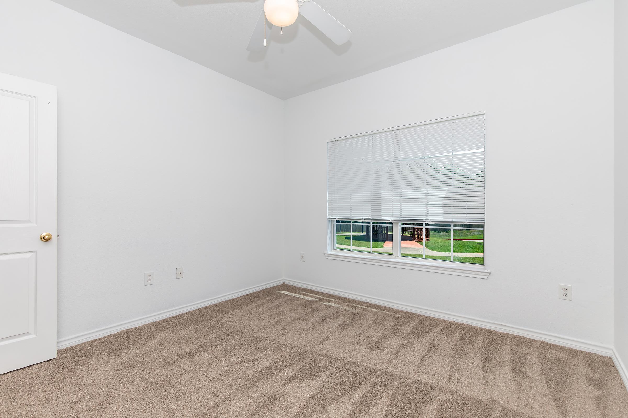 A bright, empty room featuring light-colored walls and a beige carpet. The window has white blinds and overlooks a green outdoor area. A ceiling fan is installed, and there is a white door on the left side of the image. The room has a clean, spacious feel with natural light streaming in.