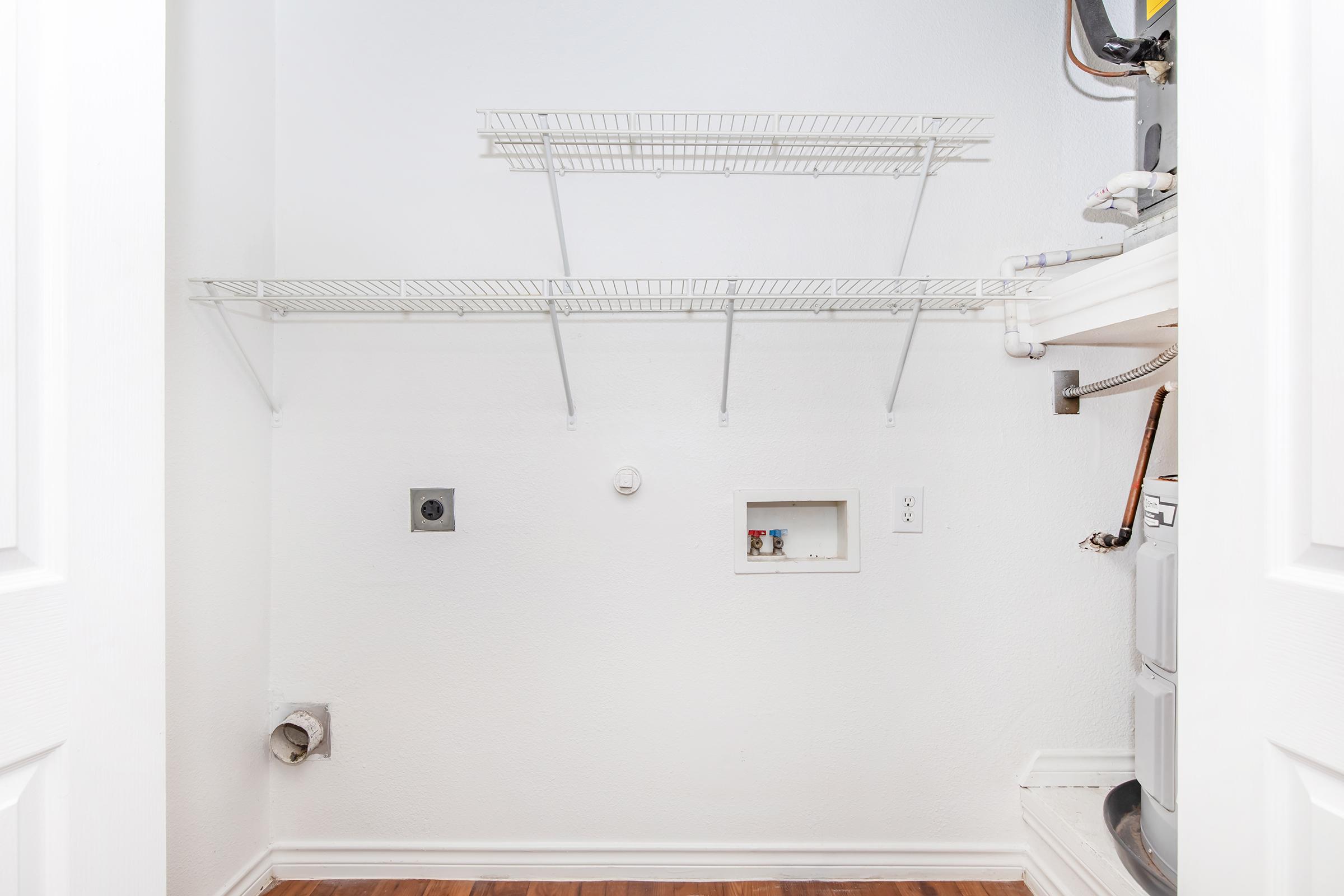 A clean and empty laundry room featuring white walls and a wooden floor. There are wire shelves installed on the wall, along with plumbing and electrical outlets visible. An area for a washing machine is indicated, but no appliances are present. The space is well-lit and organized.