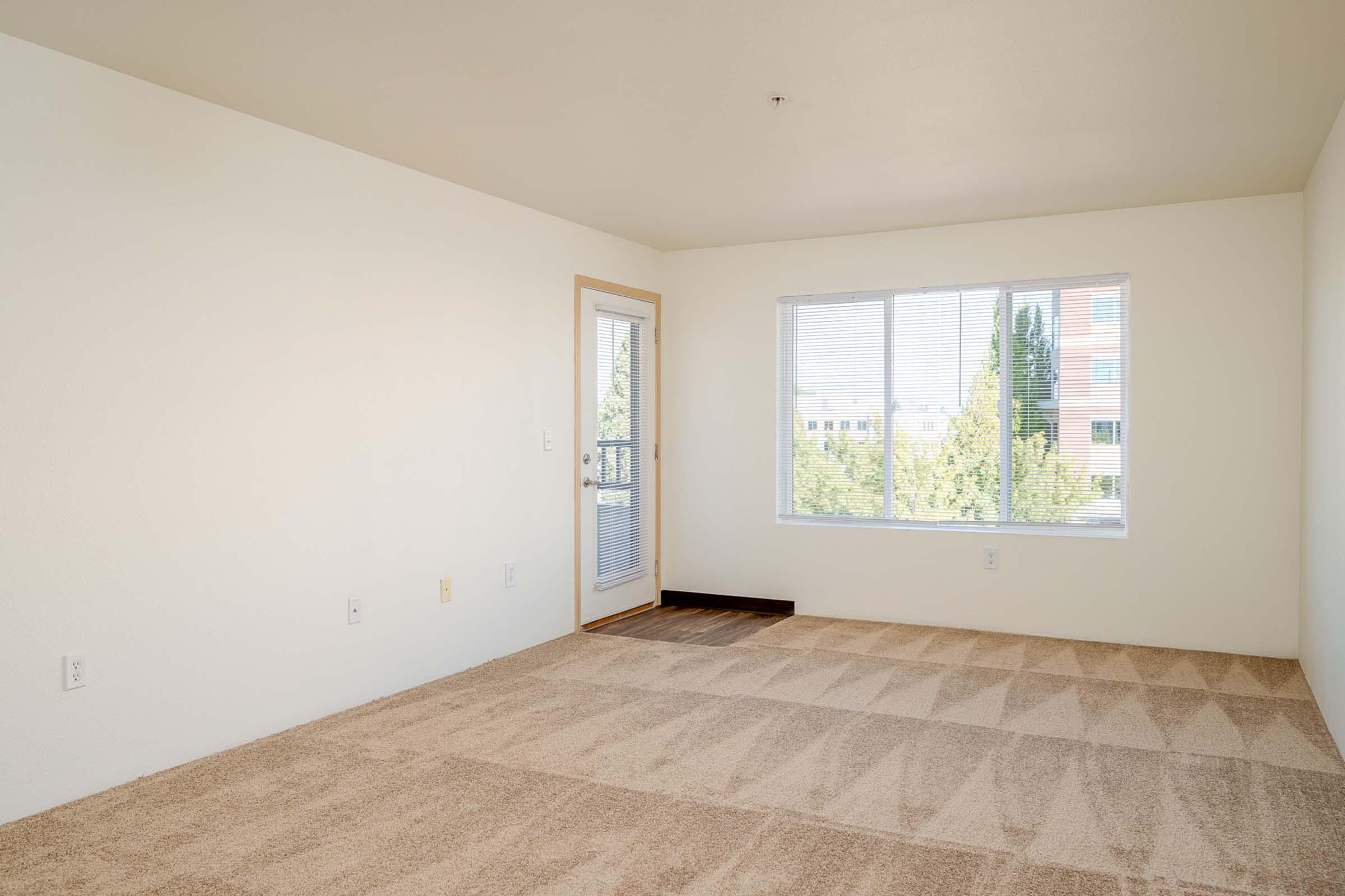 A bright, empty room with light-colored walls and a large window allowing natural light. The floor is covered in light beige carpet, and there is a small door leading to a balcony or exterior area. The overall atmosphere is spacious and inviting, ideal for personal customization.