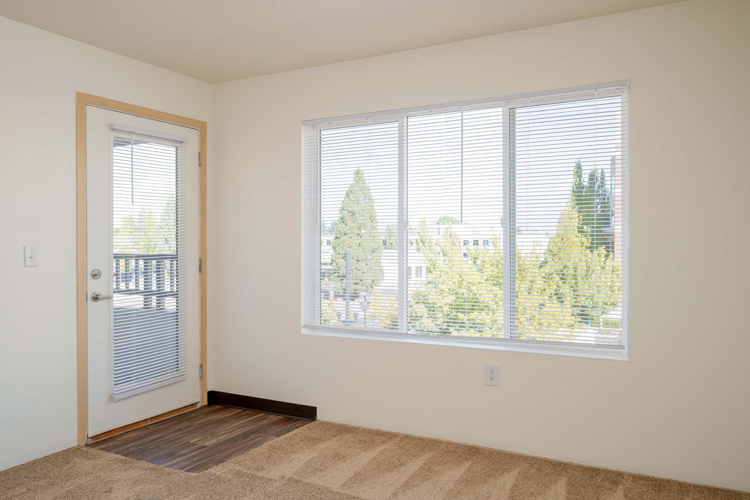 An interior view of a room with light-colored walls and a large window covered by blinds. The window offers a view of green trees outside. A sliding glass door is visible on the left, and the floor is carpeted in a neutral shade.