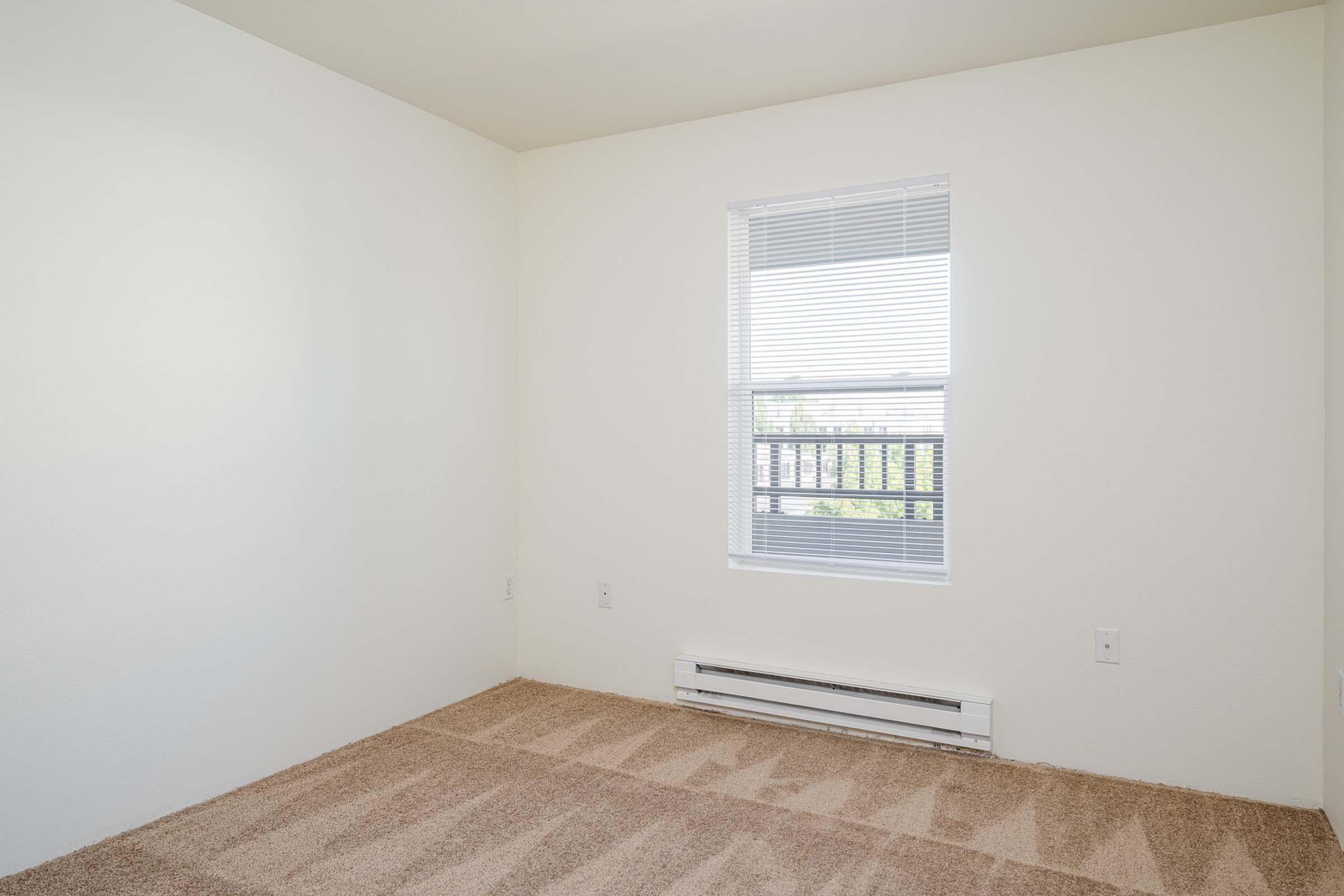 Empty room with light beige carpet and white walls. A window with blinds is visible, leading to a small balcony. The room is unadorned, creating a spacious and minimalist atmosphere.