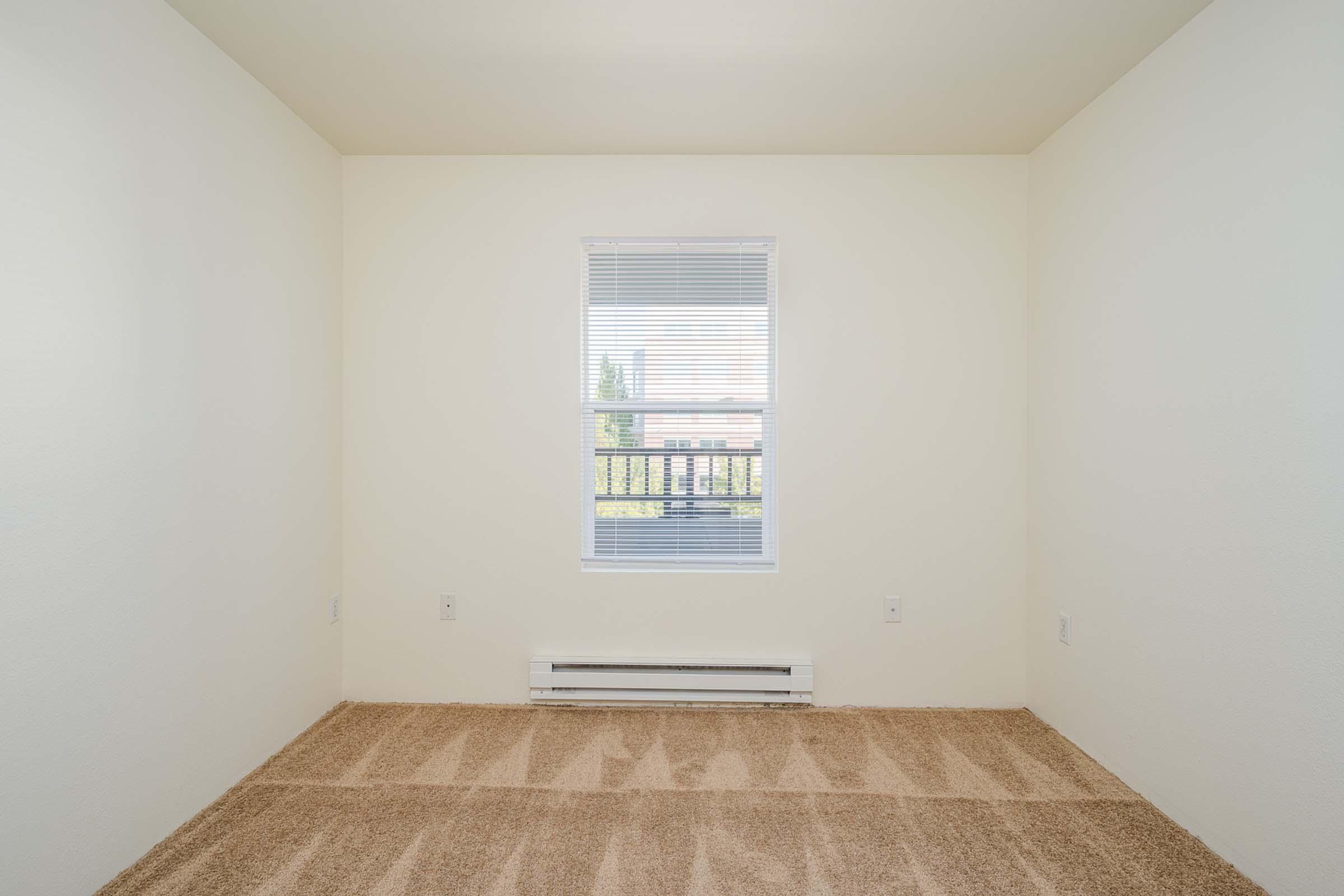 A small, empty room with beige carpet and light-colored walls. There is a window with white blinds allowing natural light to enter, and a heating unit beneath the window. The space appears clean and minimalistic, suitable for various uses.