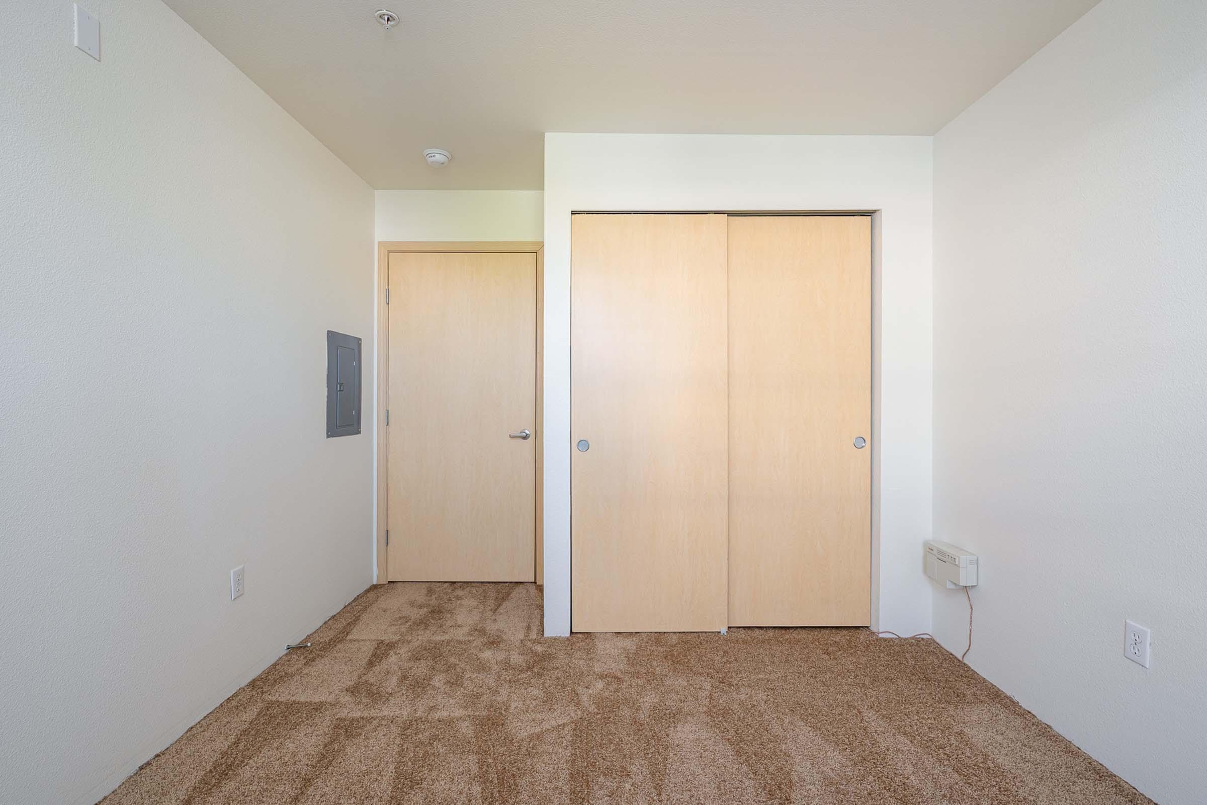 A small, empty room with light-colored walls and beige carpet. There are two wooden closet doors on the right side and a closed door to the left. A small electrical panel is mounted on the wall near the door. The room is well-lit and spacious, creating a minimalist feel.