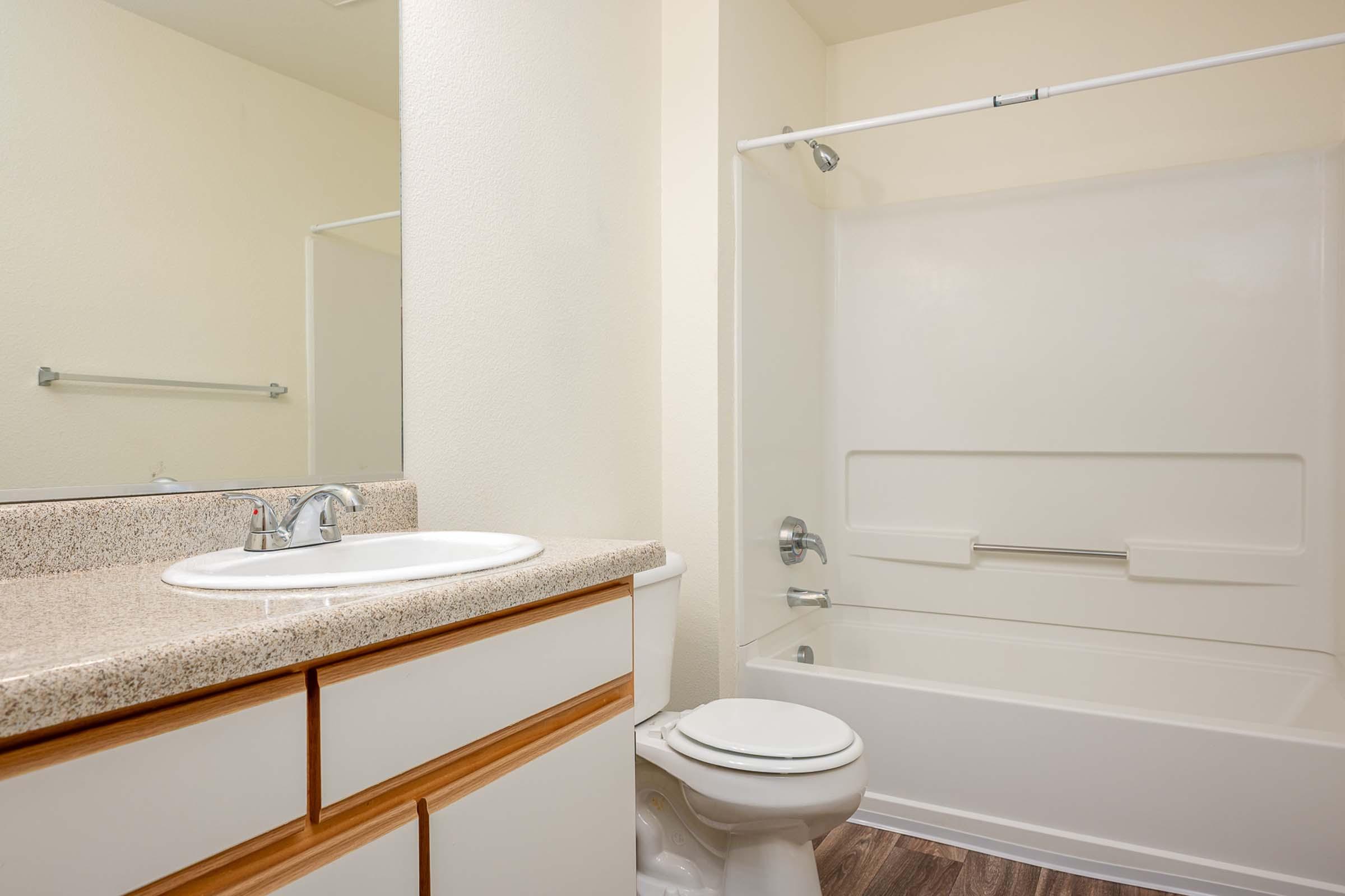 A clean and modern bathroom featuring a white bathtub with a shower curtain, a sink with a countertop, a toilet, and a large mirror. Light-colored walls and a wooden floor create a bright and spacious atmosphere.