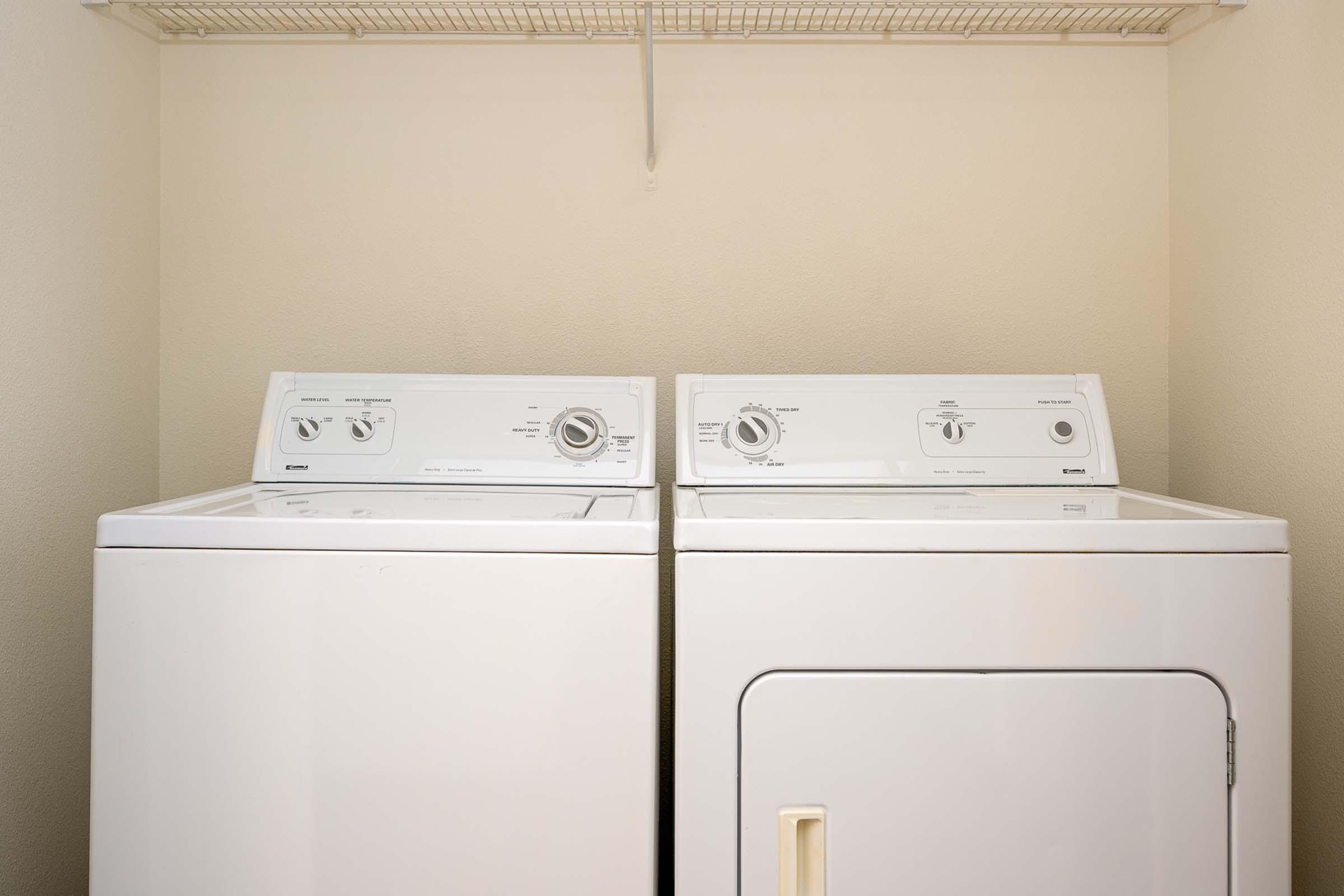A white washing machine and dryer side by side, set in a light-colored laundry room with a shelf above for storage. The appliances have control dials and are clean and modern, with a neutral wall color in the background.