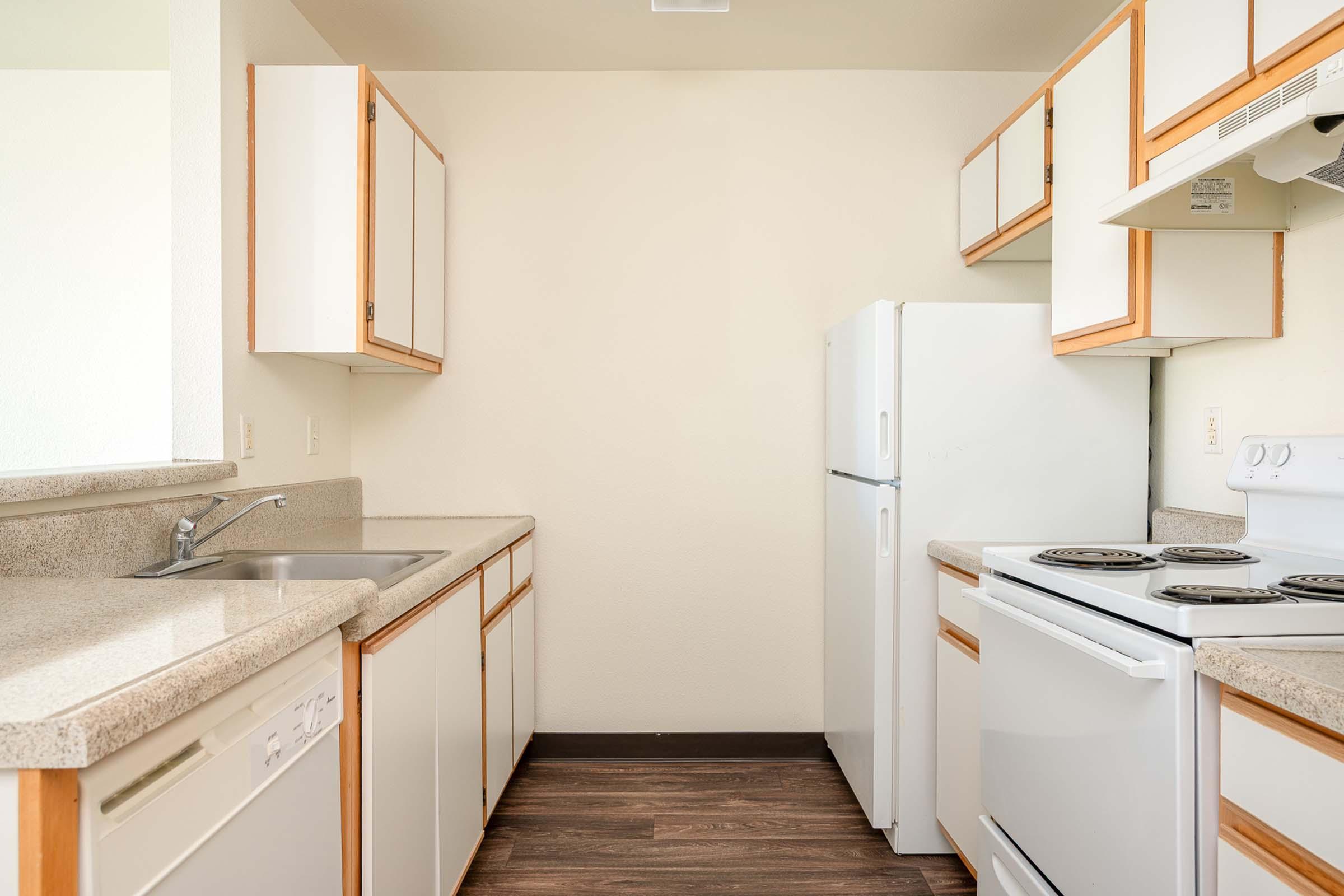 A modern kitchen with white cabinets and beige countertops. It features a stainless steel sink, a white refrigerator, a white stove with an oven, and a dishwasher. The walls are light-colored, and the flooring is dark wood laminate, creating a clean and spacious appearance.