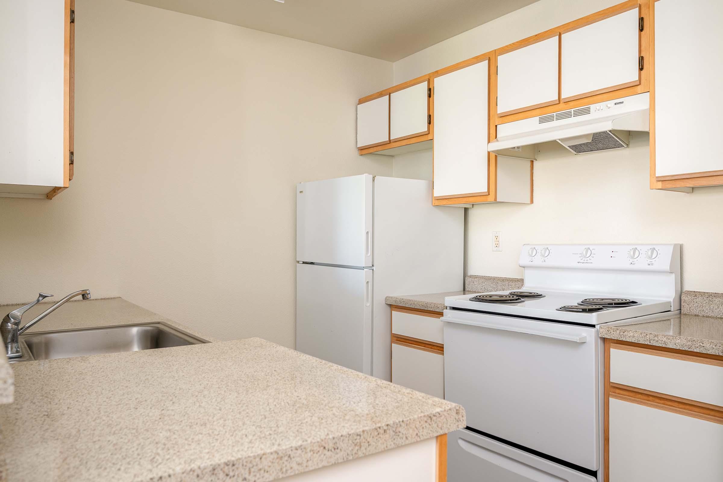 A modern kitchen featuring white cabinetry, a stainless steel sink, a white refrigerator, and a white stove with an overhead exhaust fan. The countertops are beige with a speckled design, creating a clean and bright atmosphere.