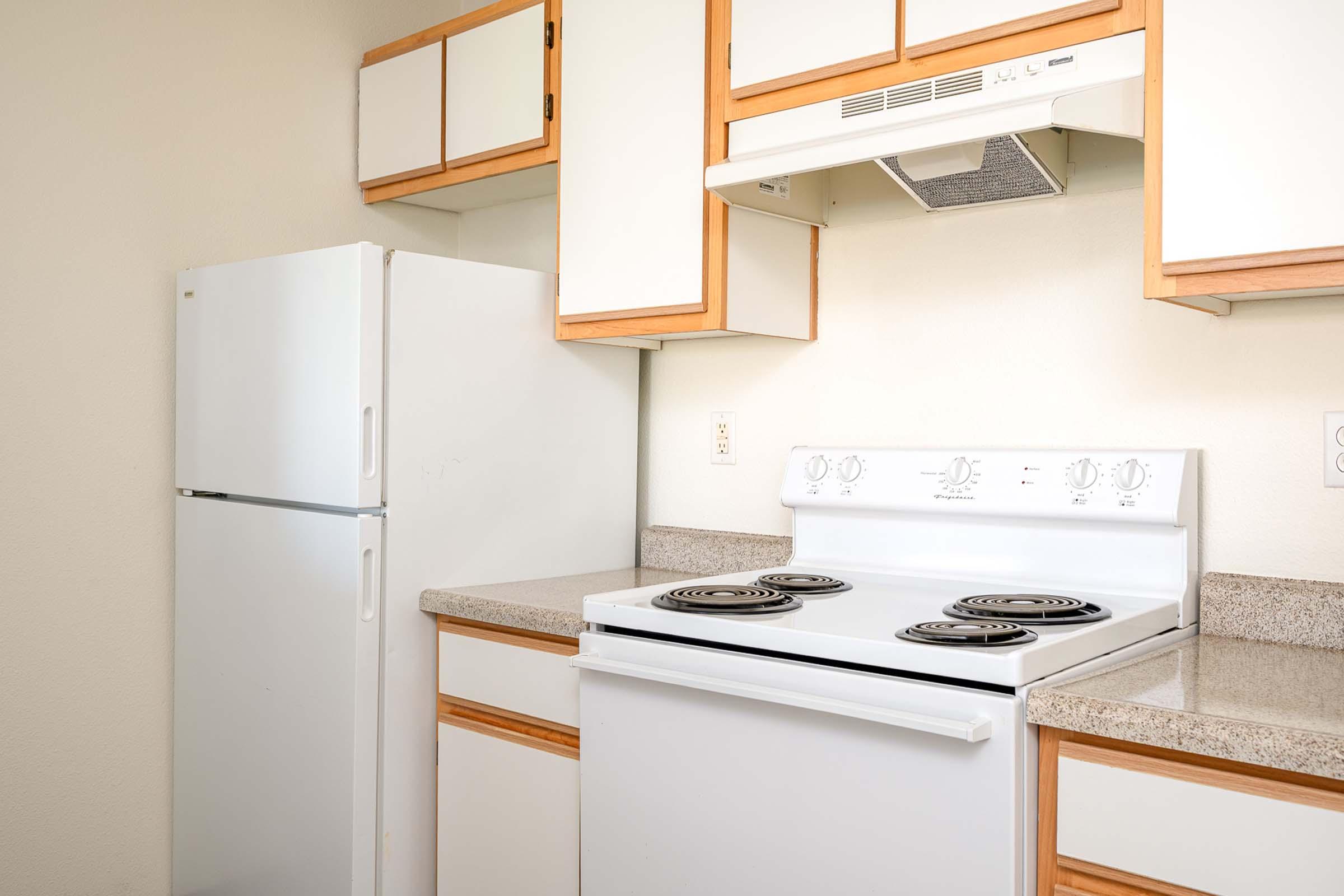 A kitchen with a white refrigerator and a white stove with four burners. There are wooden and white cabinets above the countertop, and a range hood installed above the stove. The countertop is made of beige granite. The walls are painted a light color.
