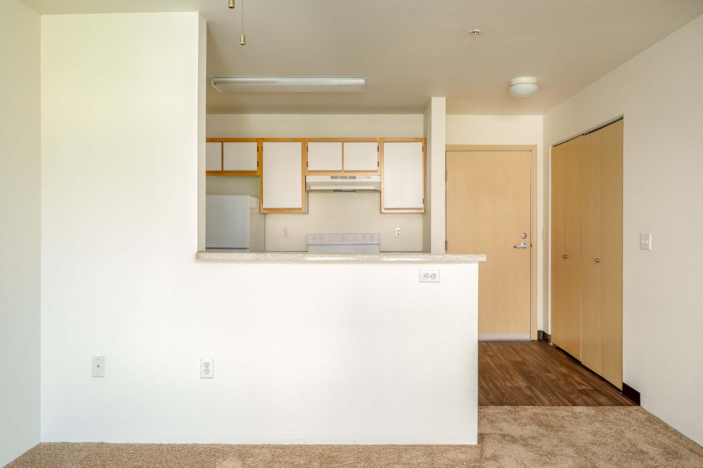 An interior view of a small kitchen area with a bar counter, featuring beige cabinets and a white stove. The adjacent door leads to another room, and the floor has light carpet, while the walls appear painted in neutral tones.