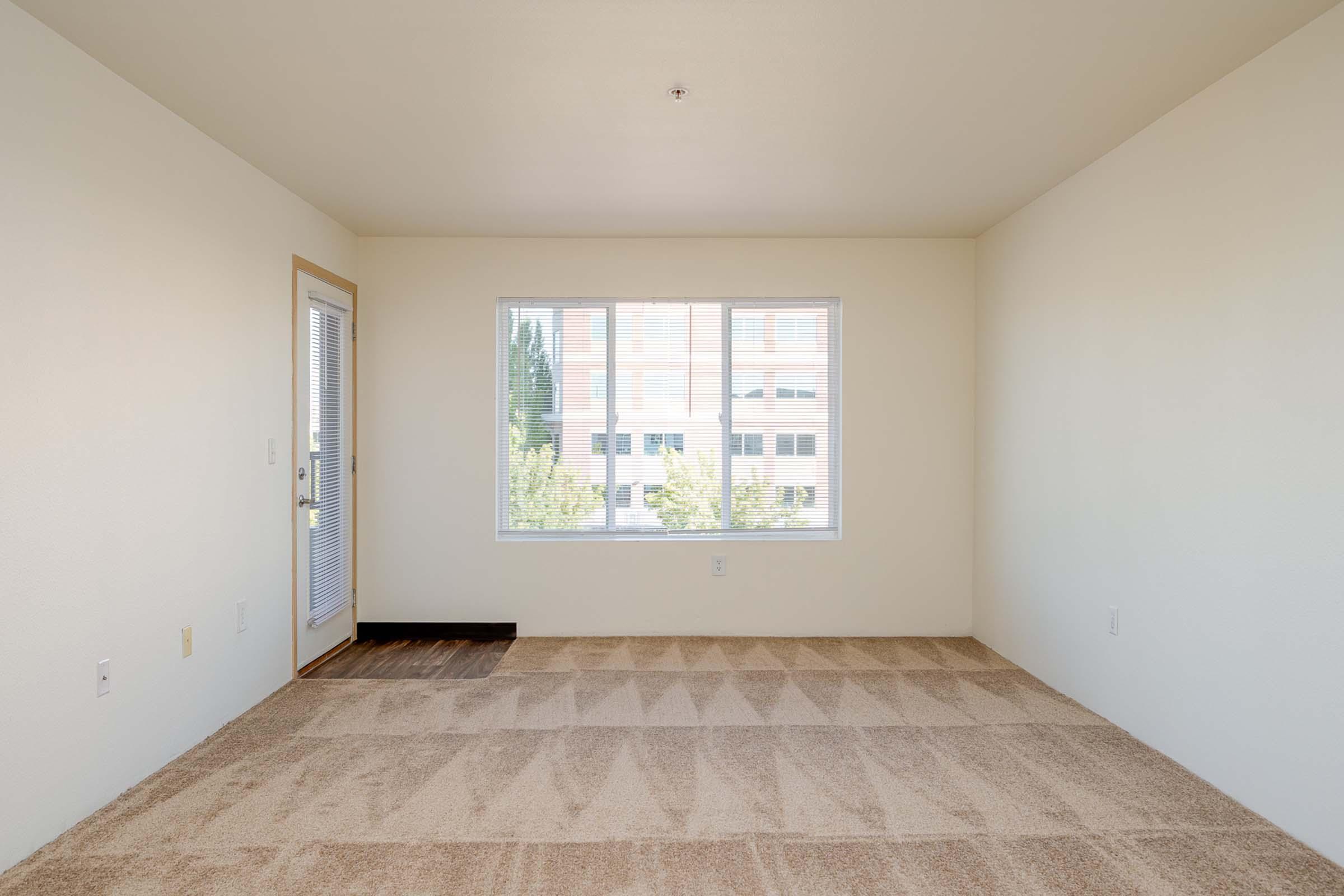 A bright, empty room with beige carpet and plain cream-colored walls. A large window provides natural light, facing a view of trees outside. A door on the left leads to a balcony or patio. The overall atmosphere is spacious and minimalistic, ideal for customization.