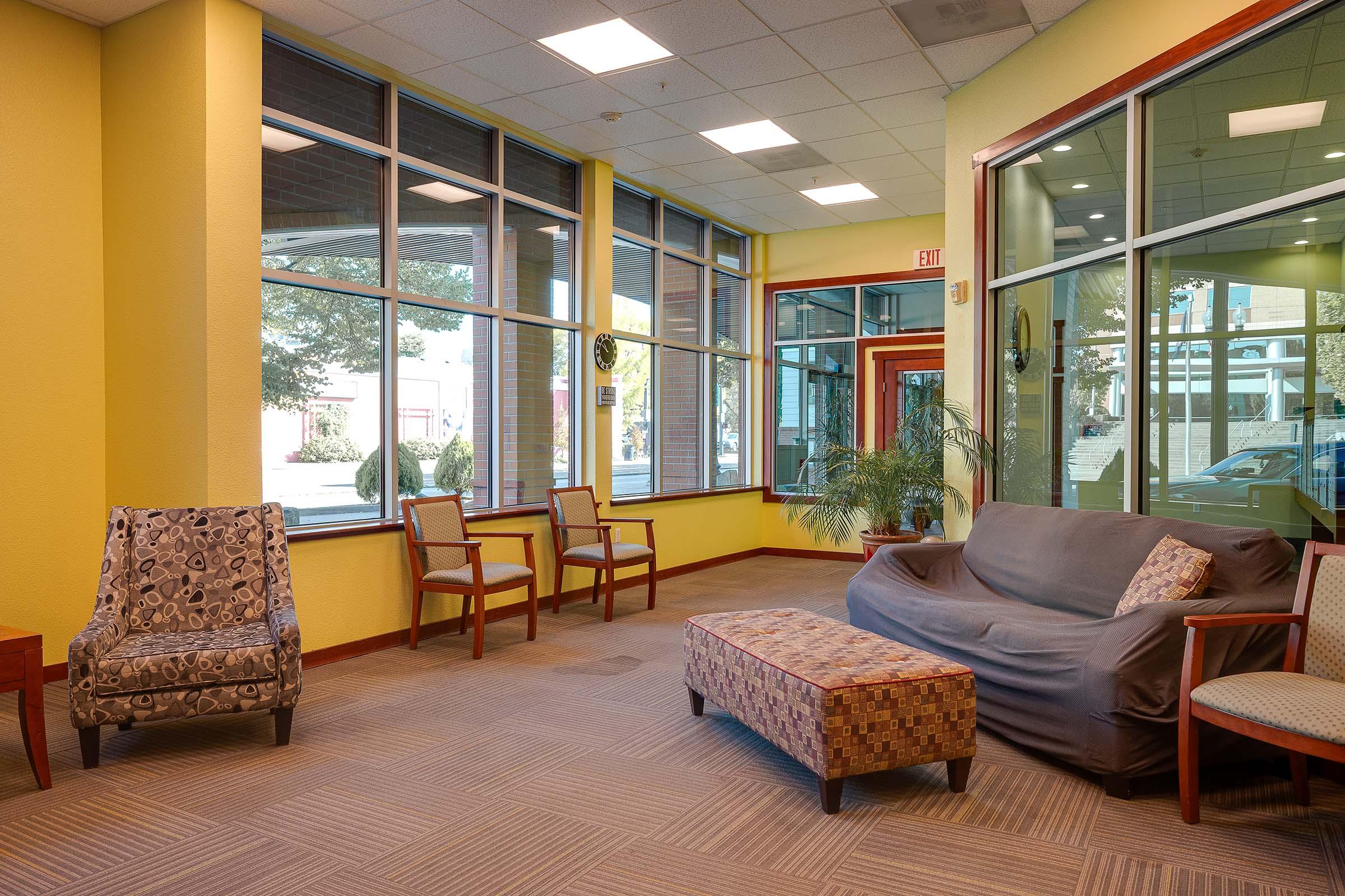 A bright and inviting lounge area featuring a gray couch, patterned chairs, and a coffee table. Large windows allow natural light to fill the space, with a potted plant adding a touch of greenery. The walls are painted a vibrant yellow, creating a warm atmosphere.