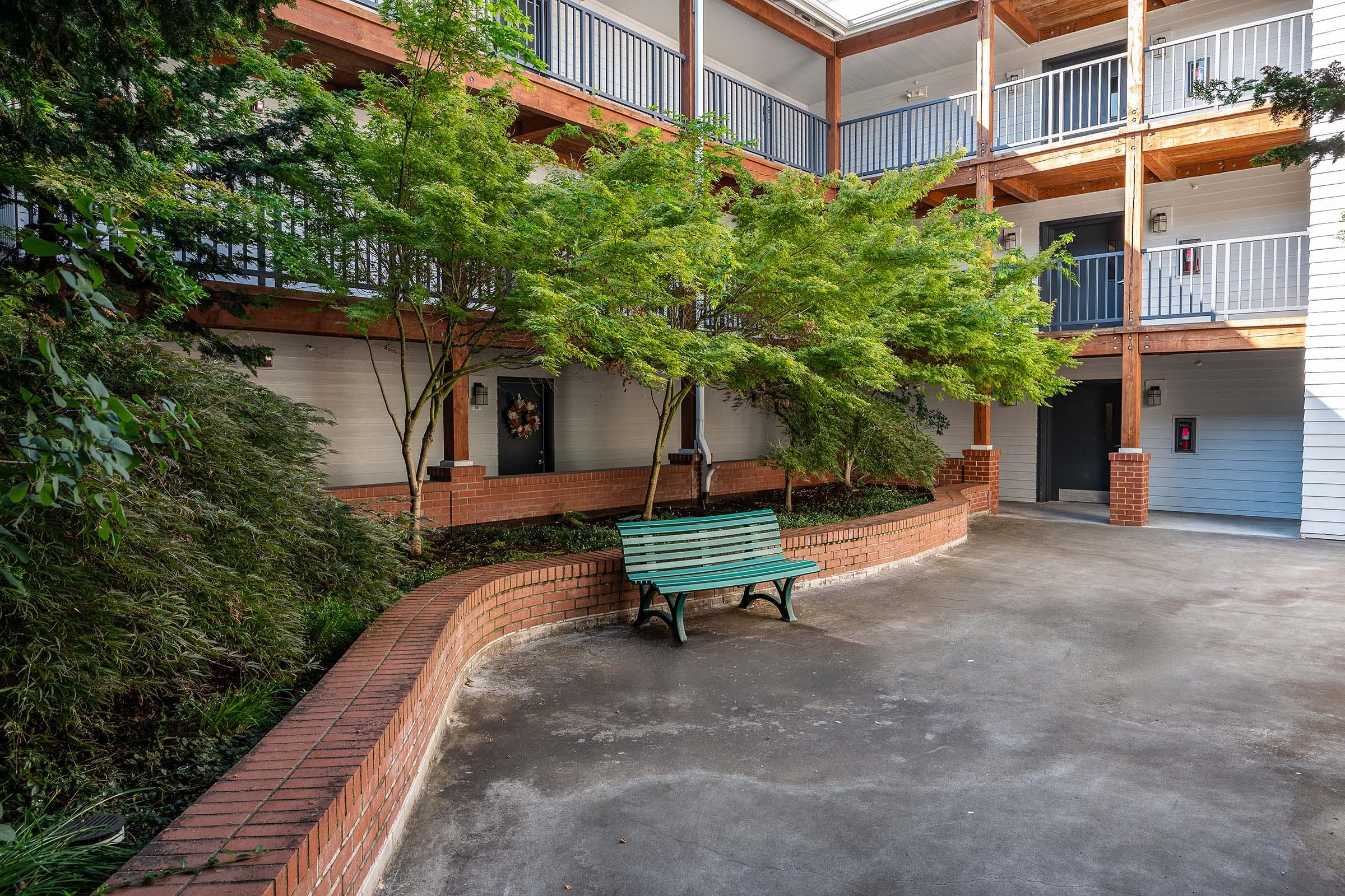 A serene courtyard with a green bench surrounded by brick landscaping. Lush greenery and trees provide shade, while balconies line the upper levels of the surrounding building. The area features a concrete pathway, creating a peaceful and inviting atmosphere.