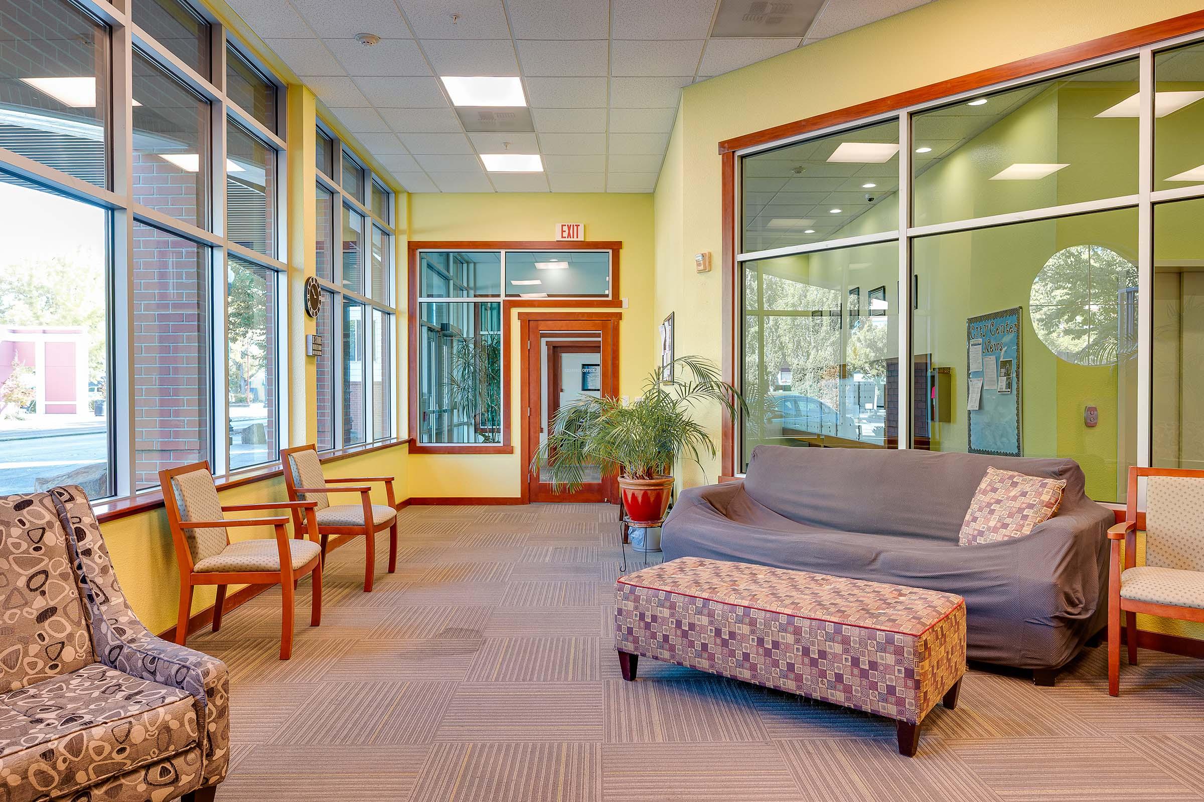 A bright, welcoming indoor space featuring a seating area with a gray couch, patterned ottoman, and various chairs. Large windows allow natural light, and plants add a touch of greenery. The walls are painted in warm yellow, enhancing the inviting atmosphere.