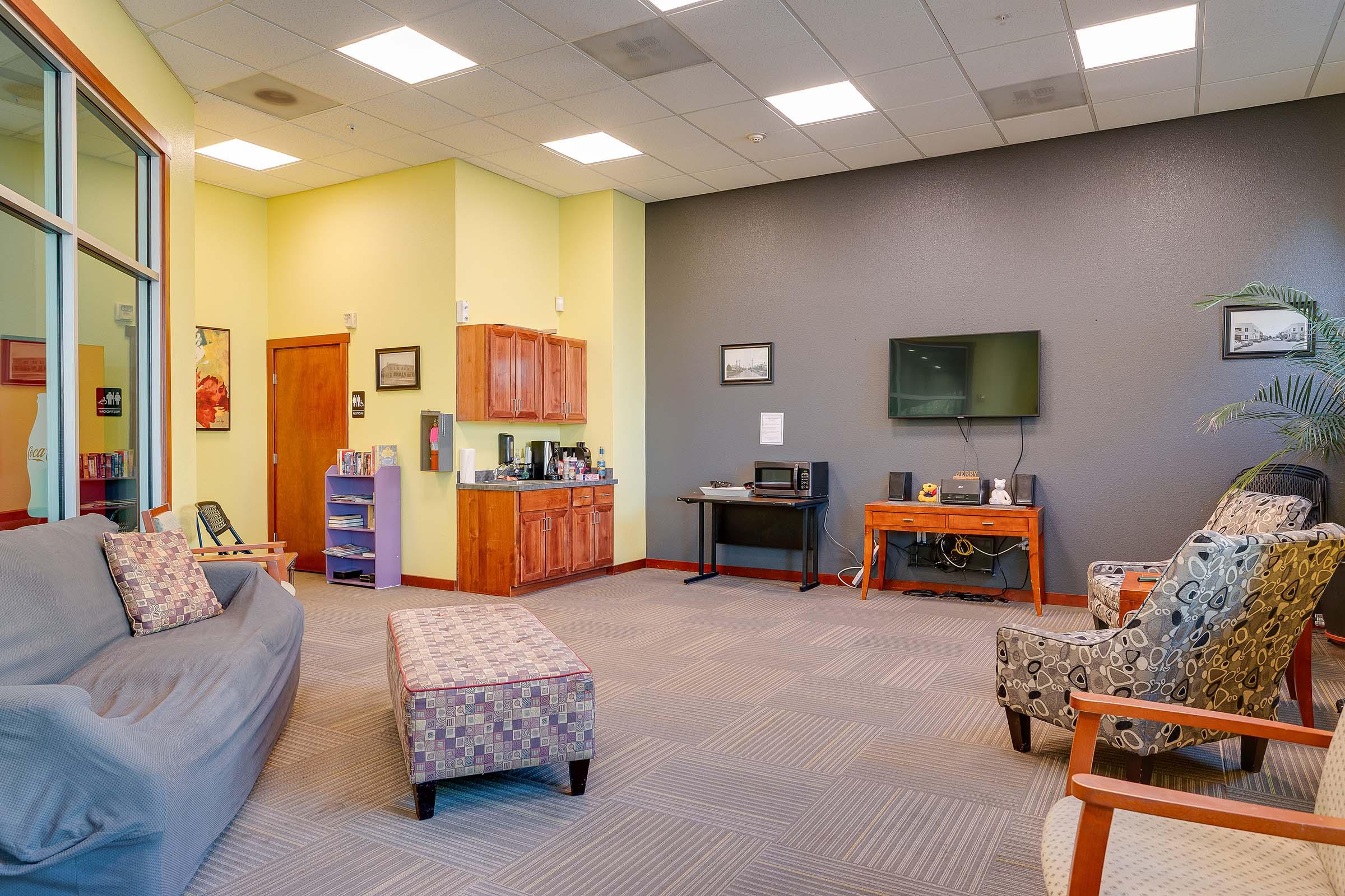 A spacious, well-lit lounge area featuring a gray sofa, patterned ottoman, and wooden chairs. A small kitchenette with cabinets and a coffee station is visible. A television mounted on the wall is present, along with a few framed pictures. The walls are painted in light yellow and gray tones.