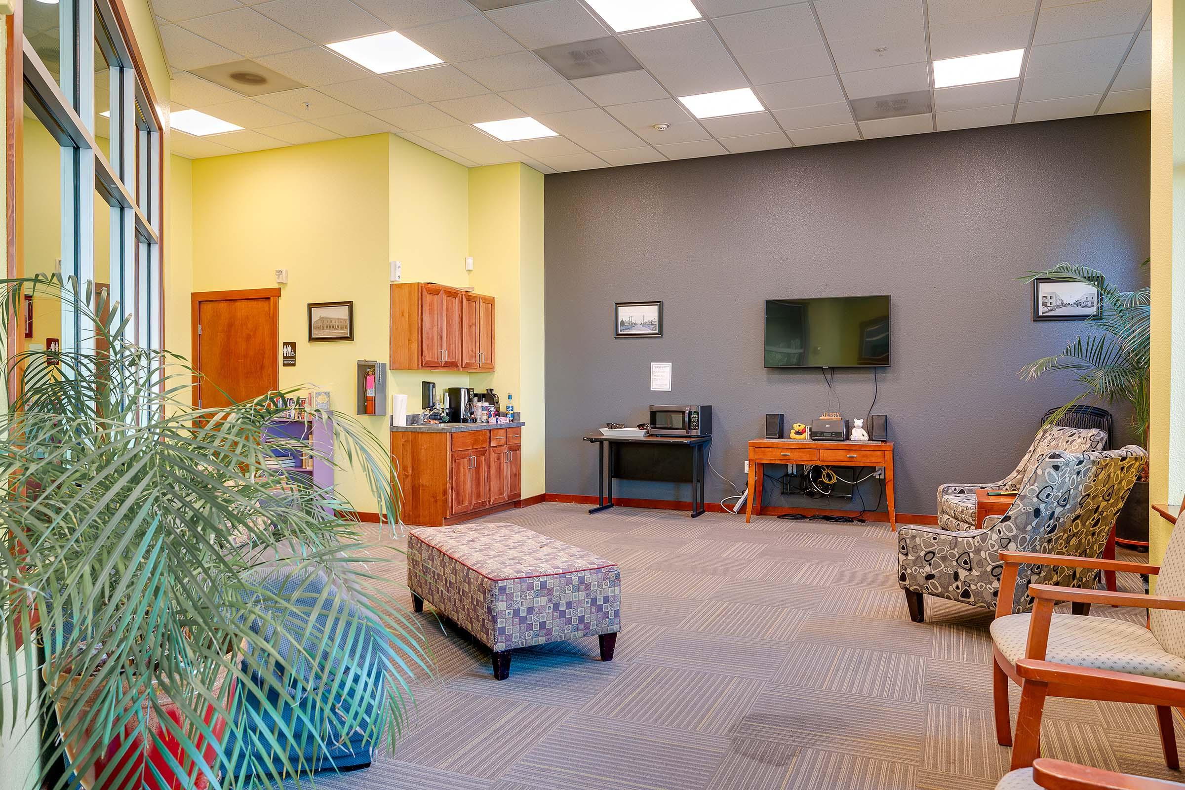 A bright, welcoming waiting area featuring a colorful, patterned bench, two comfortable chairs, a television on the wall, and a kitchenette with cabinets and appliances. Green plants add a touch of nature to the space, which has large windows letting in natural light.