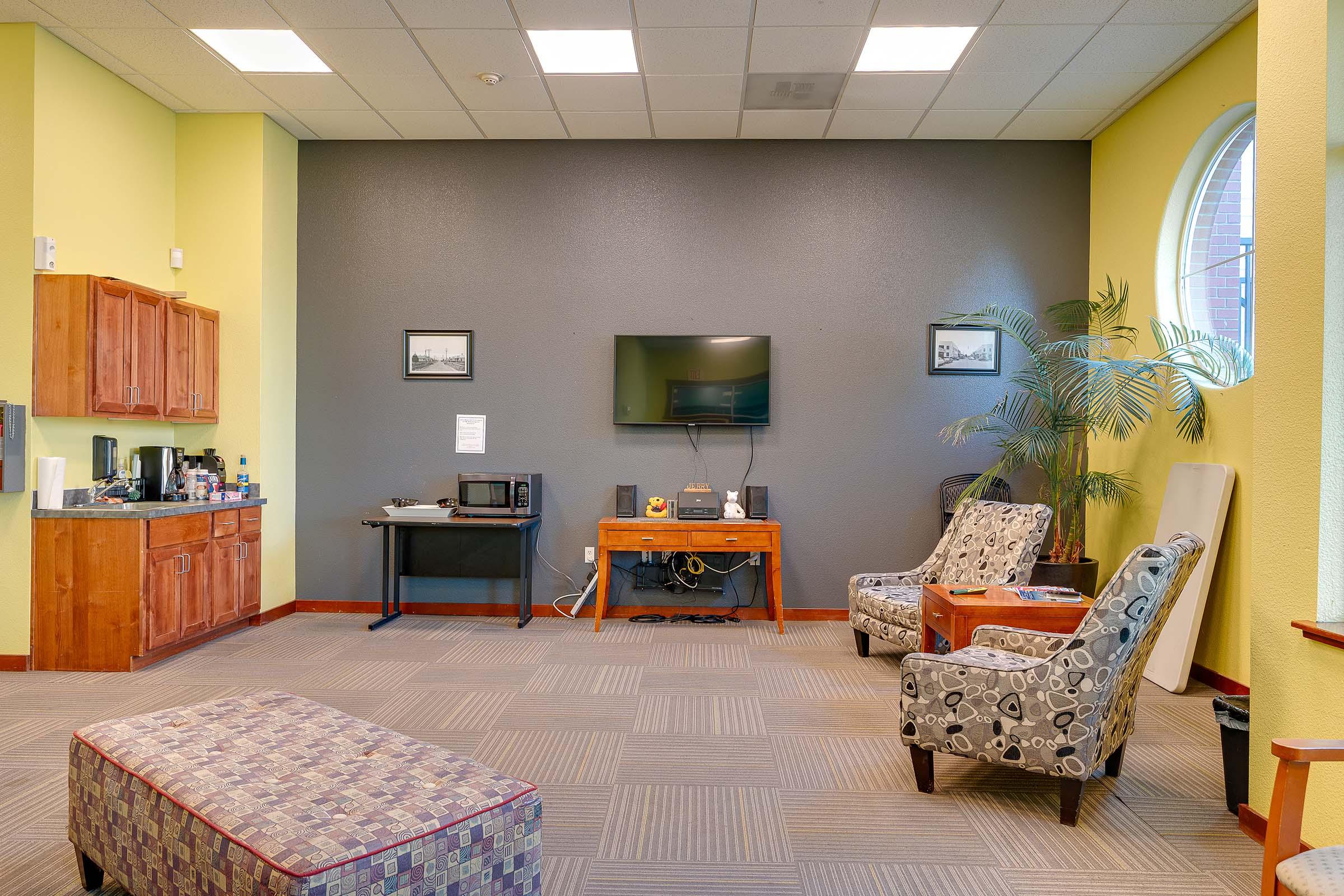 Cozy lounge area featuring a dark gray wall, a television mounted above a wooden console table, and two patterned armchairs. There is a coffee station with cabinets, a microwave, and a decorative plant. A patterned ottoman sits in the center of the room, complemented by tiled flooring and soft lighting.