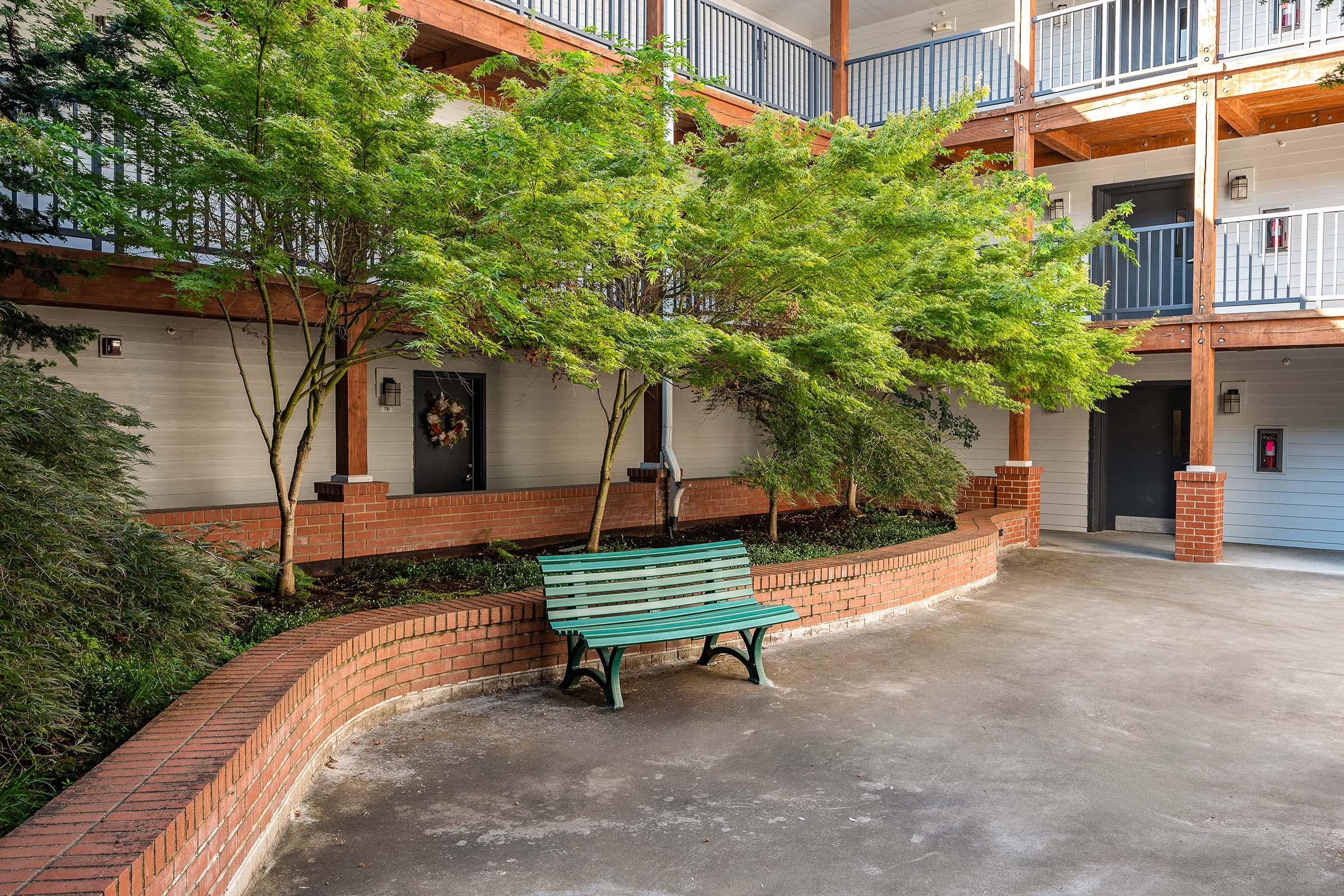 A peaceful courtyard featuring a green wooden bench surrounded by landscaped greenery, including small trees and brick planters. The area is enclosed by two levels of balconies and has access doors leading into the buildings in the background.