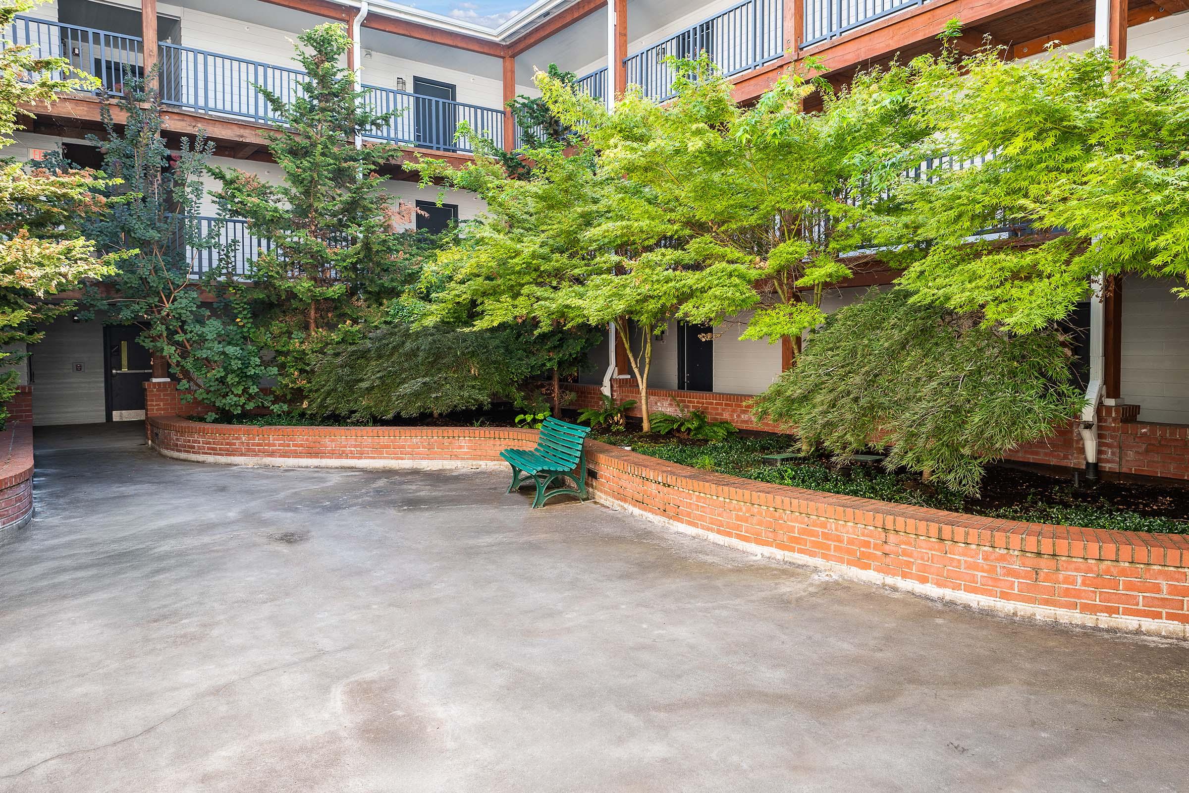 A serene courtyard with lush greenery, including small trees and shrubs, surrounding a curved brick wall. A green bench sits invitingly on the concrete path, while the background features multiple floors of a building with balconies. The setting is bright and welcoming, perfect for relaxation.